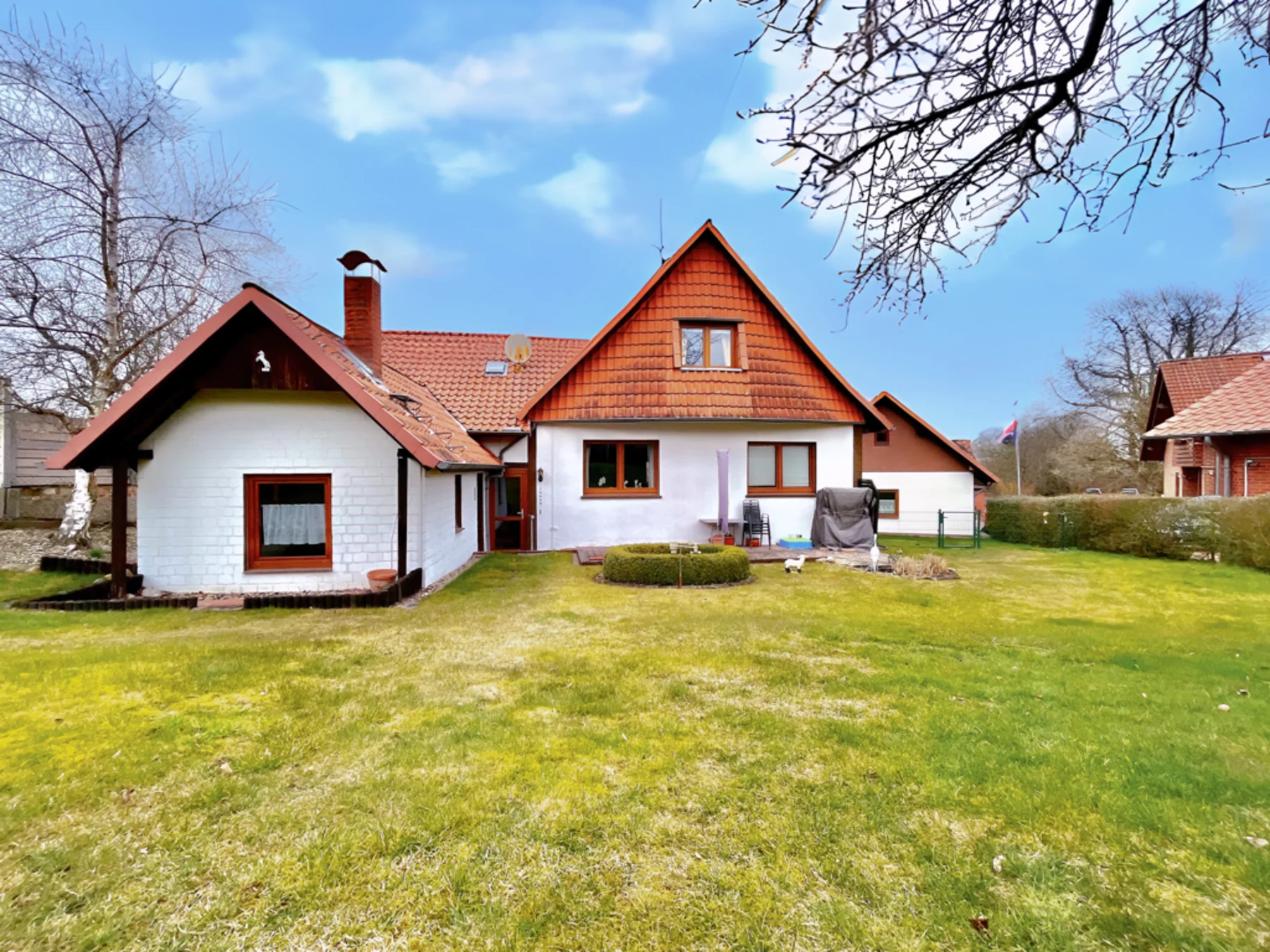 Einfamilienhaus mit Garten, rotem Dach und Terrasse, umgeben von Bäumen und Rasenfläche.