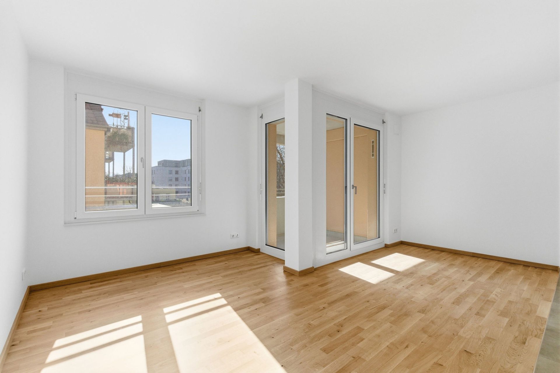 Heller leerer Raum mit Parkettboden, zwei Fenstern und einer verglasten Tür zu einem Balkon.