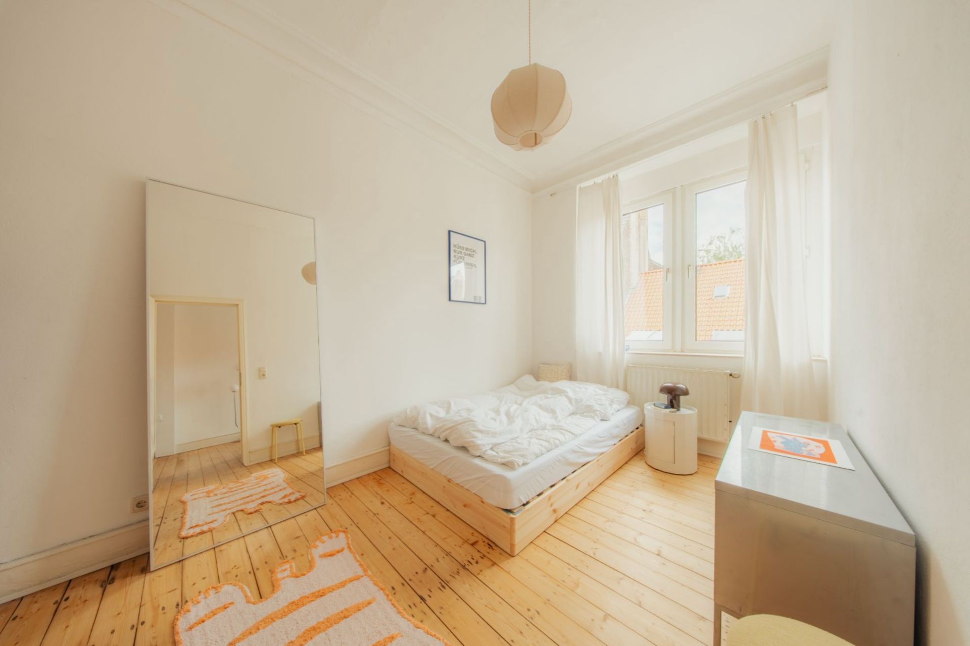 Helles Schlafzimmer mit Holzfußboden, Bett, großem Spiegel und drei Fenstern.
