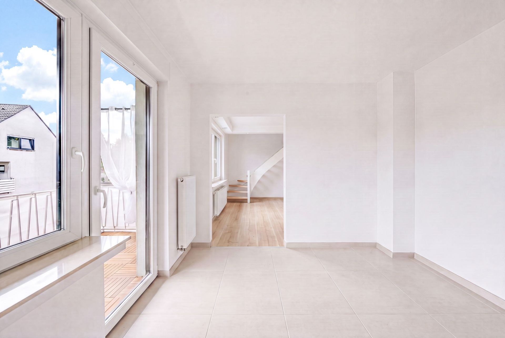 Helles, leeres Zimmer mit großen Fenstern, Fliesenboden und Blick auf Balkon und benachbarte Häuser.