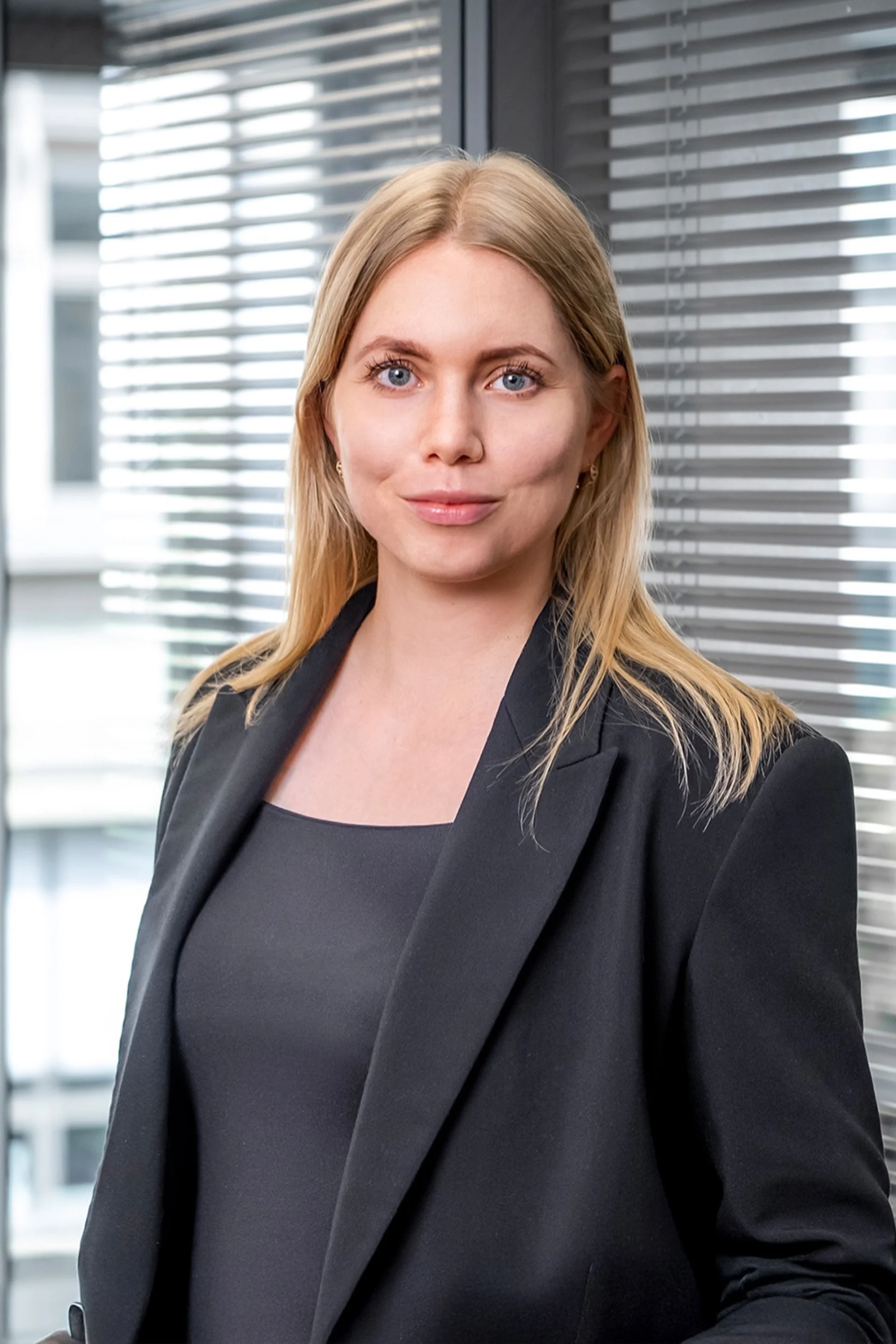 Frau mit langen blonden Haaren und schwarzem Blazer vor einem Fenster mit Jalousien.