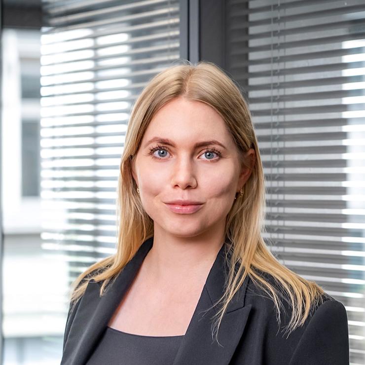 Frau mit langen blonden Haaren und schwarzem Blazer vor einem Fenster mit Jalousien.
