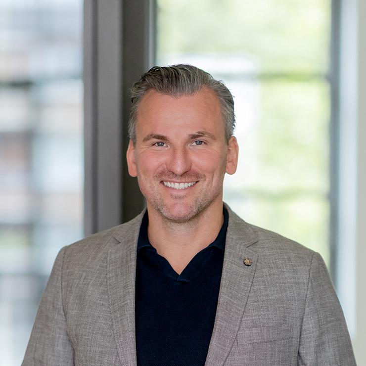 Ein lächelnder Mann im grauen Sakko steht vor einem Fenster in einem Innenraum.