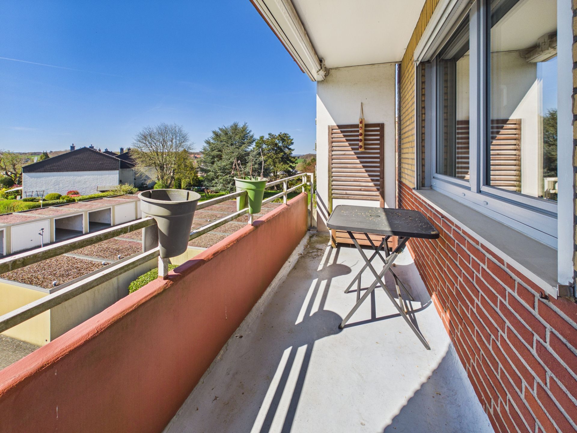 Balkon mit Tisch, Stuhl und Blumentöpfen, Blick auf Nachbarhäuser und Bäume bei Sonnenschein.
