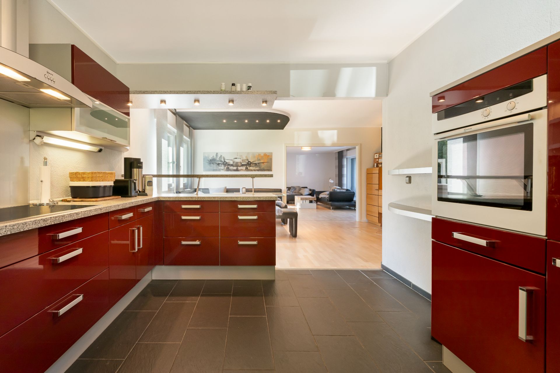 Moderne Küche mit roten Hochglanzschränken und Einbaugeräten, angrenzend an ein offenes Wohnzimmer.