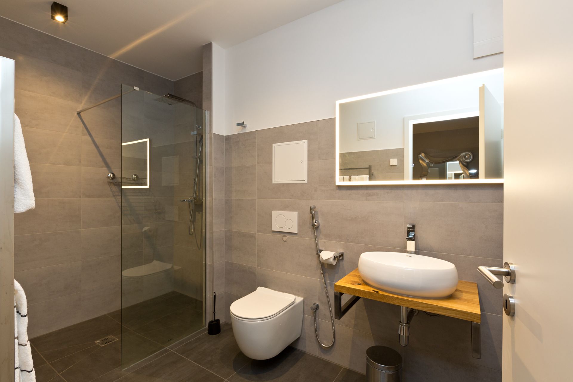 Modernes Badezimmer mit Dusche, Toilette und Waschbecken auf Holzplatte, beleuchteter Spiegel.