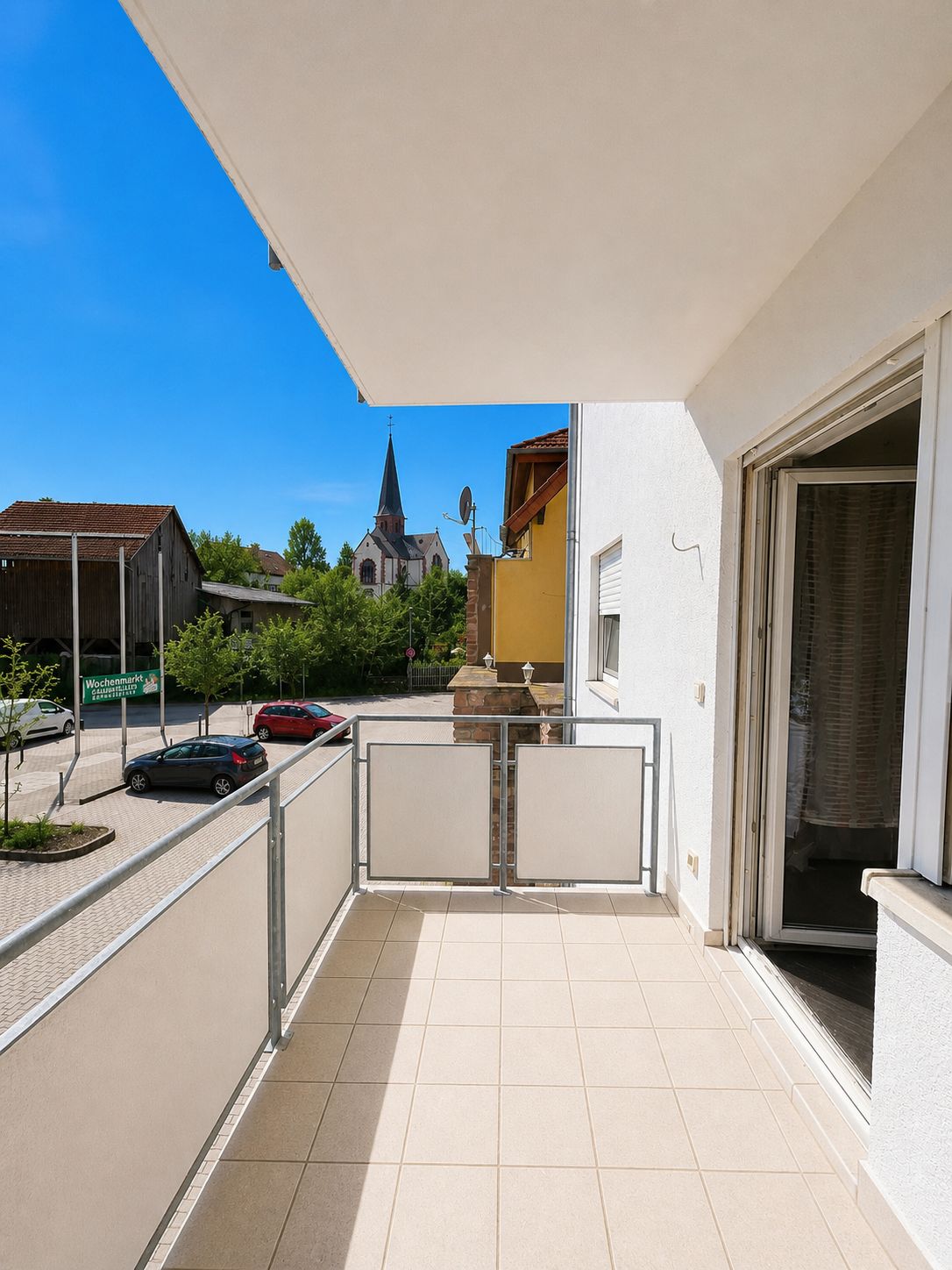 Balkon mit Fliesenboden, Blick auf Straße, Bäume und Kirche im Hintergrund.