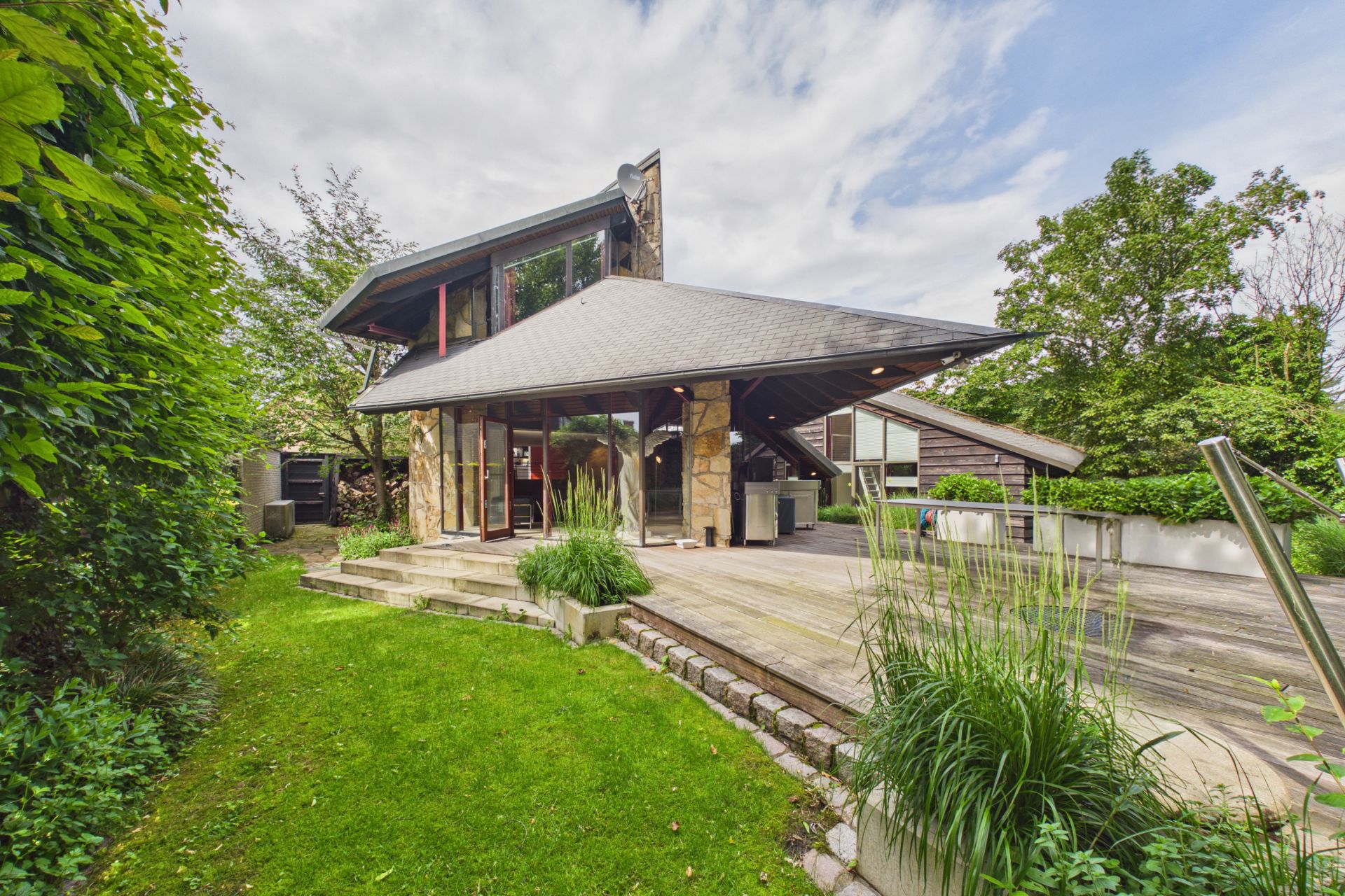 Modernes Wohnhaus mit großer Holzterrasse, Steintreppen und viel Grün im Garten.