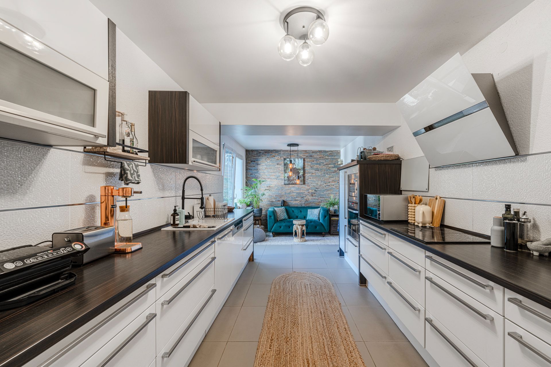 Moderne Küche mit weißen Schränken, dunklen Arbeitsplatten und Blick auf ein Wohnzimmer mit grünem Sofa.