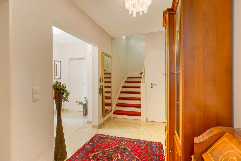 Heller Flur mit rotem Teppichläufer, Treppe, Holzmöbeln und Kronleuchter.