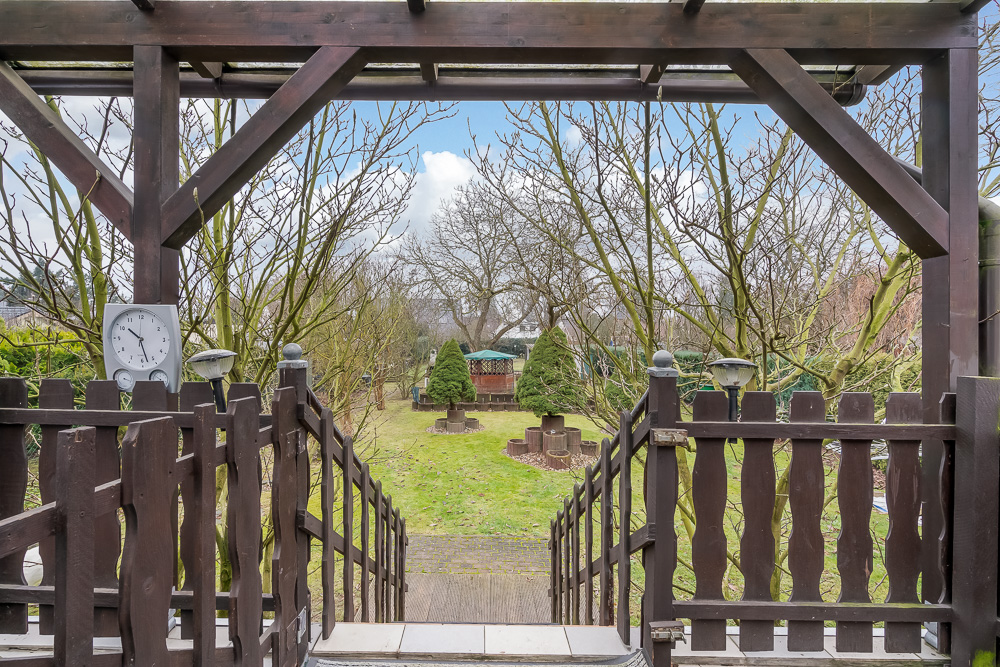 Blick von einer Holzterrasse auf einen Garten mit Bäumen, Zaun und Pavillon.
