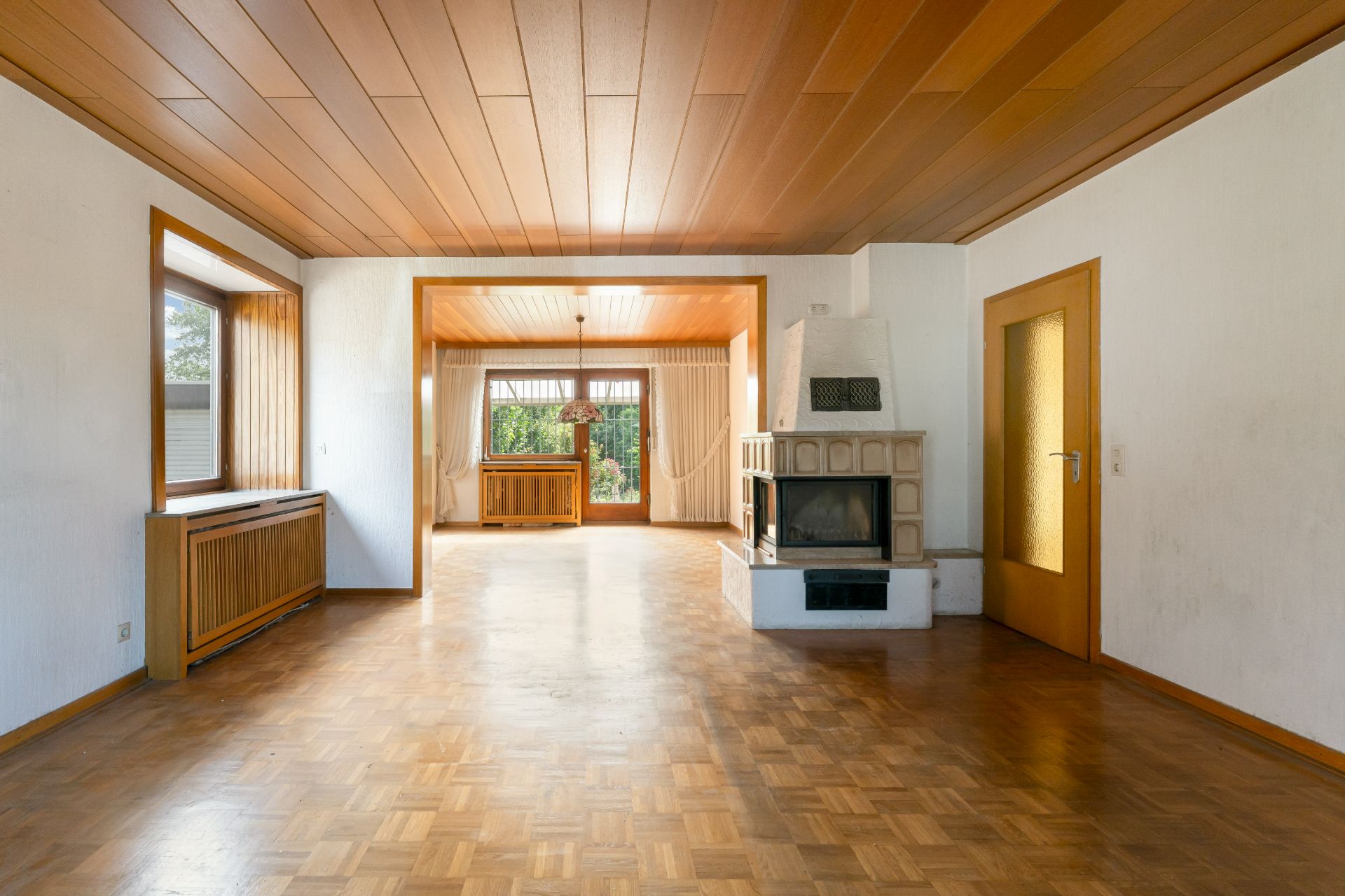 Wohnzimmer mit Holzboden, Kamin, Holzdecke und großen Fenstern, Blick auf grünen Garten.