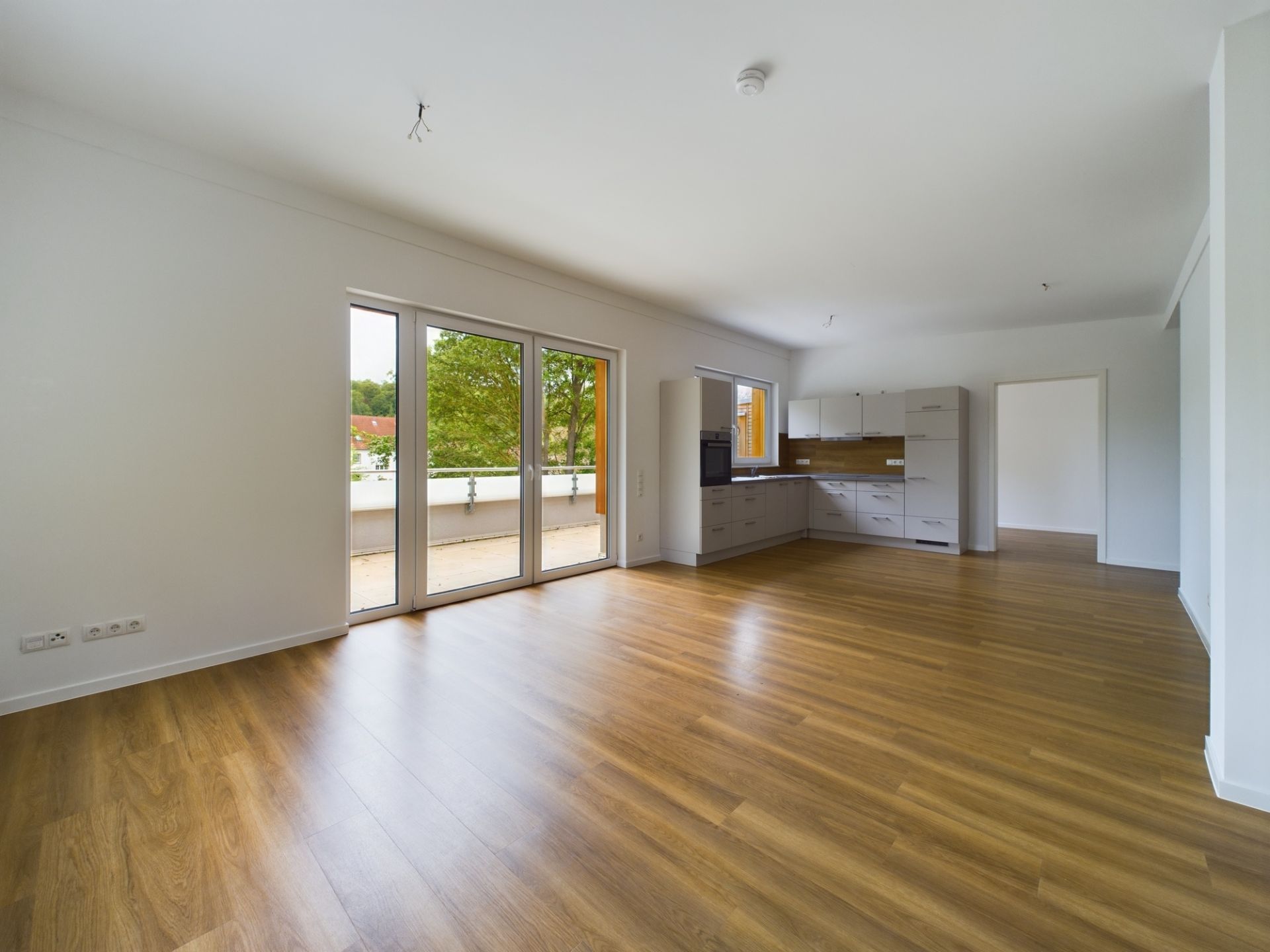 Helles, leeres Wohnzimmer mit offener Einbauküche und Parkettboden, große Fenster zur Terrasse.