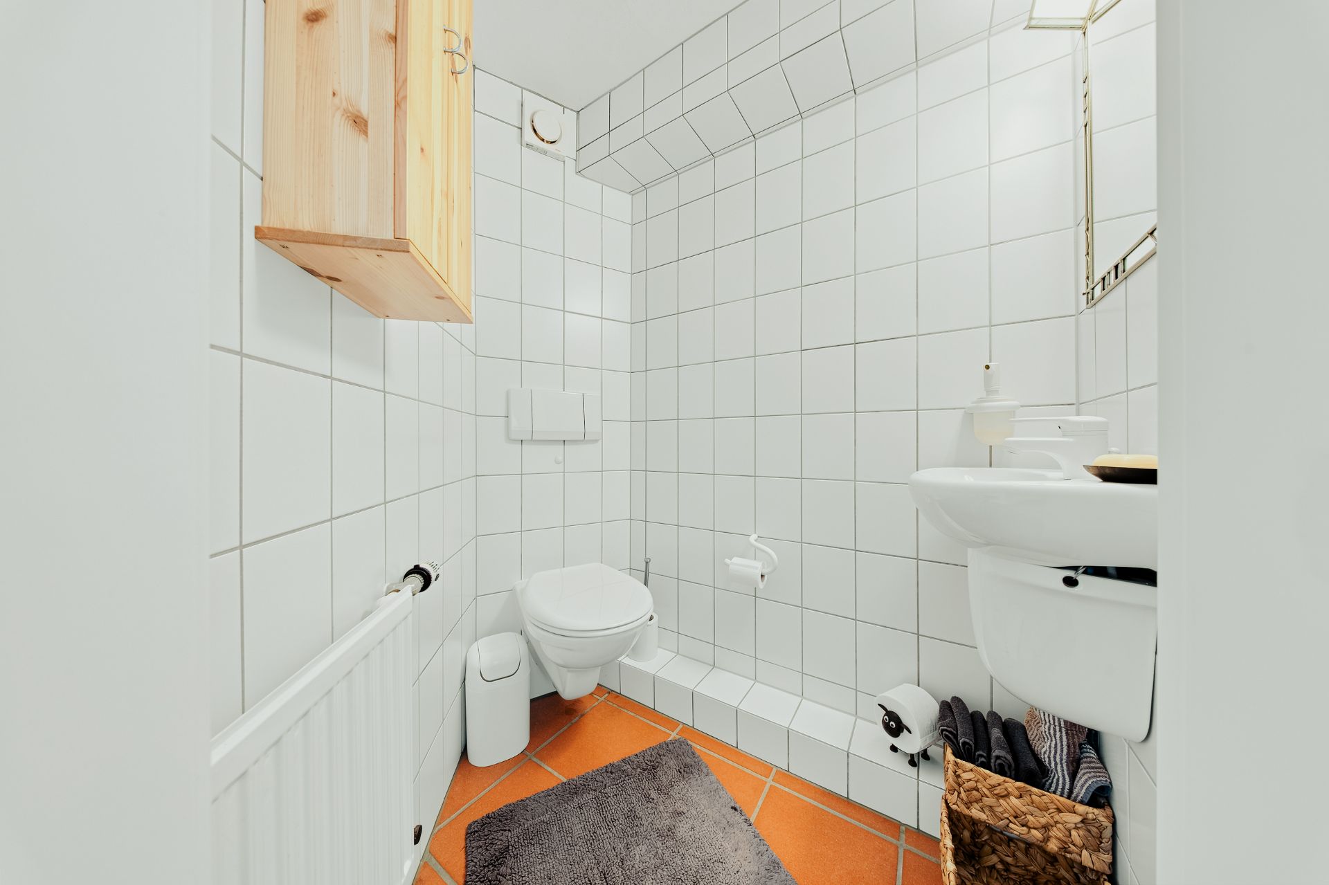 Helles Badezimmer mit WC, Waschbecken, Spiegel und Fliesenwänden, braunem Schrank und Korb.