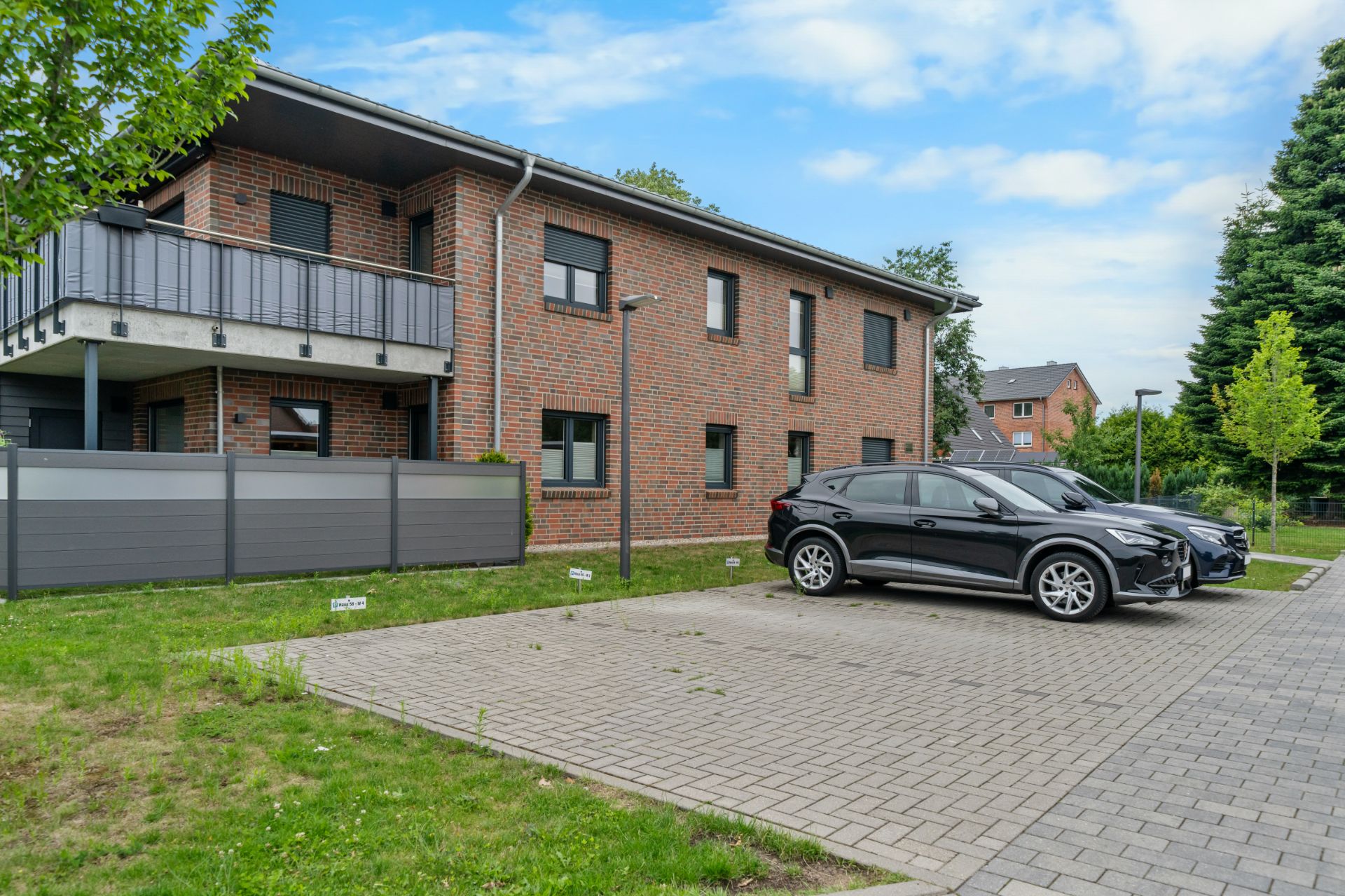 Backsteinwohnhaus mit Balkon und zwei geparkten Autos auf einem gepflasterten Parkplatz.