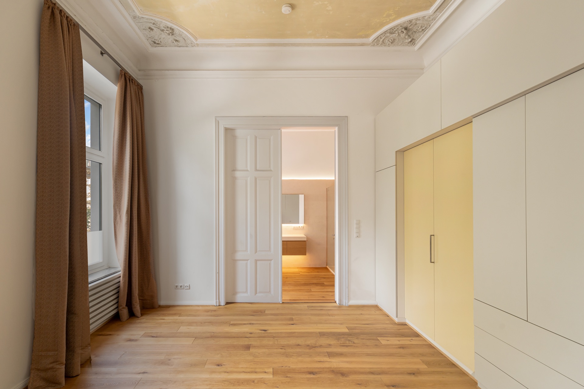 Heller Raum mit Holzfußboden, großem Fenster, Einbauschrank und Blick ins Bad.