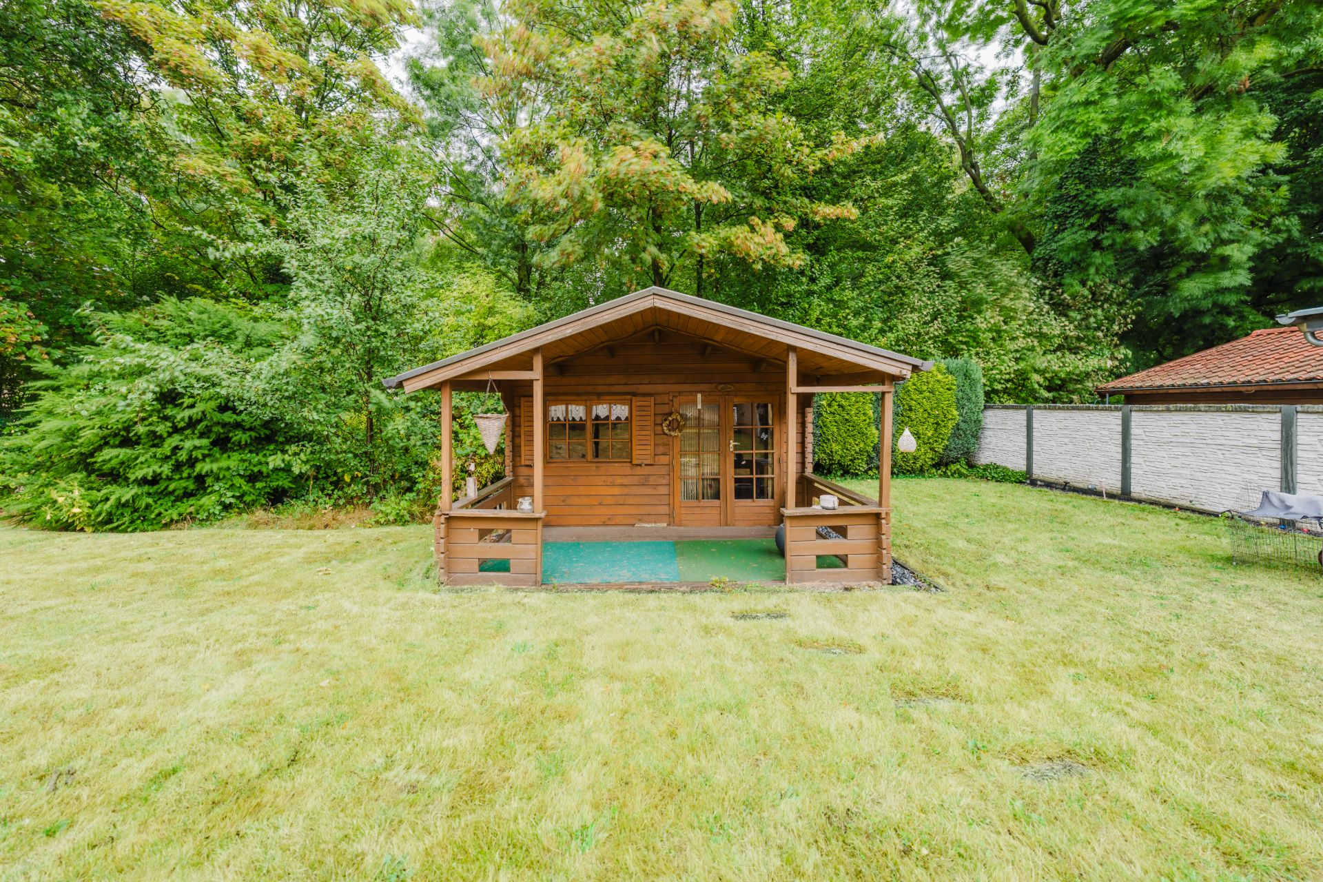 Kleines Holzhaus mit Veranda steht auf einer Wiese, umgeben von Bäumen und Sträuchern.