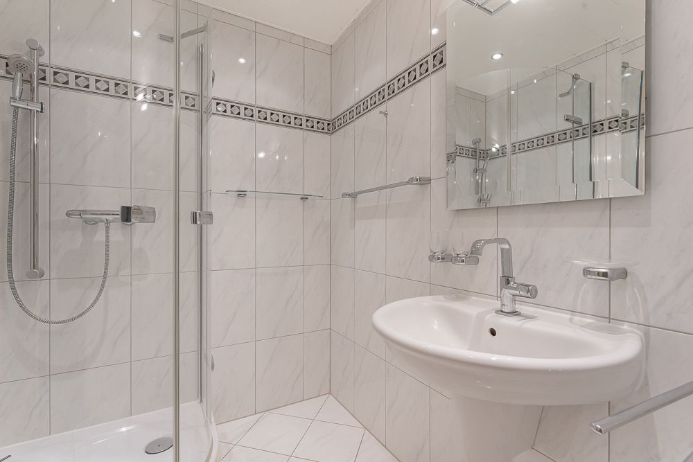 Modernes Badezimmer mit Dusche, Waschbecken und großem Spiegel, weiße Fliesen mit Musterbordüre.