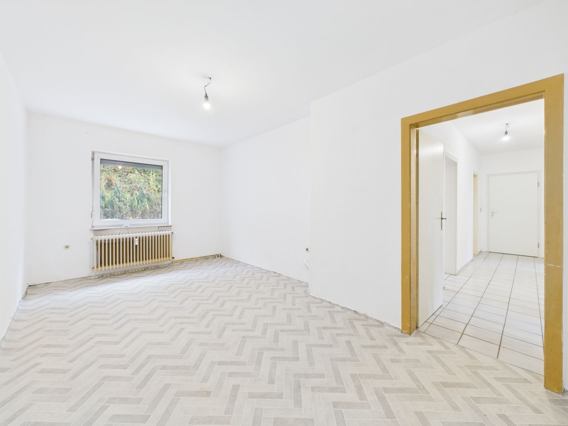 Leeres, helles Zimmer mit großem Fenster, Heizkörper und gemustertem Boden, Blick in einen Flur.