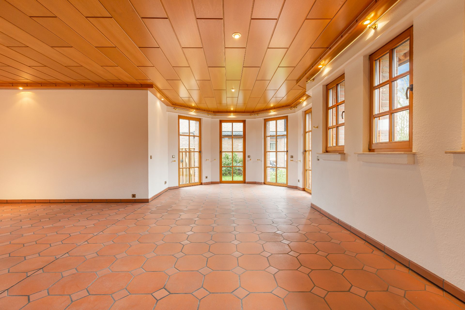 Heller, leerer Raum mit Terrakottafliesen, Holzbalkendecke und großen Fenstern.