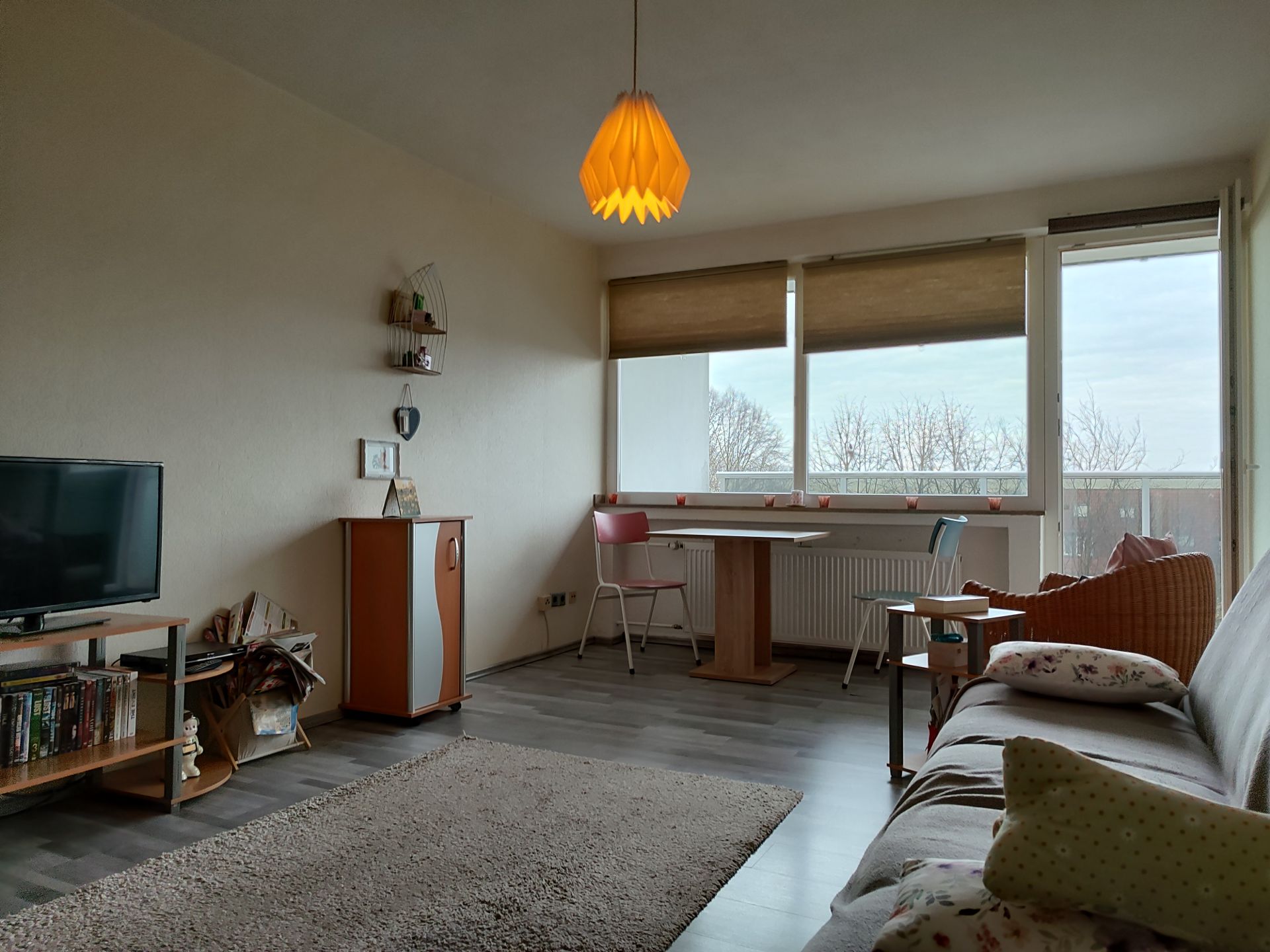 Helles Wohnzimmer mit Sofa, TV, Esstisch, Stühlen und großem Fenster mit Blick nach draußen.