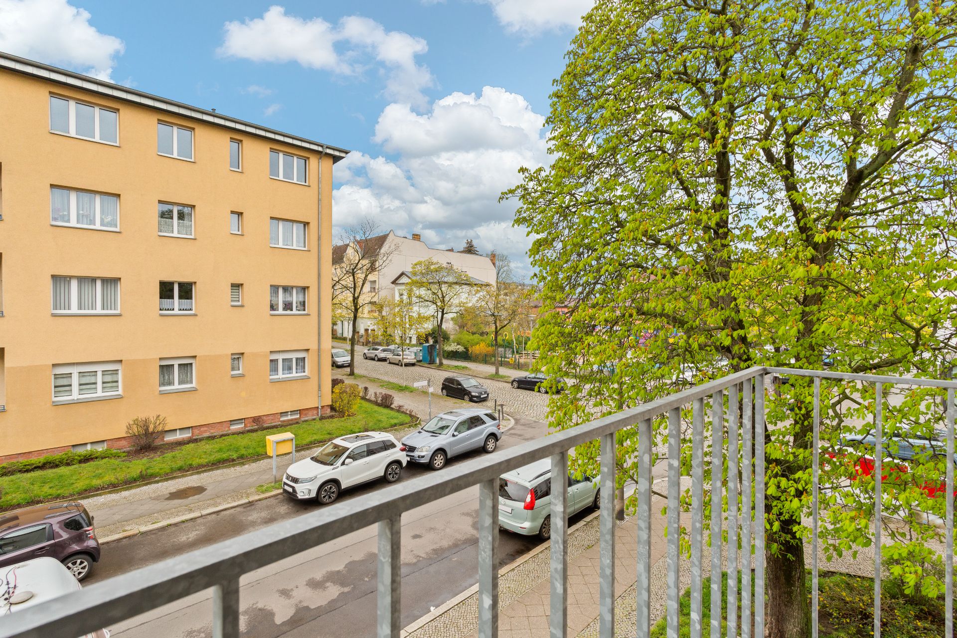 Blick von einem Balkon auf eine Straße mit parkenden Autos, Bäumen und einem gelben Wohngebäude.