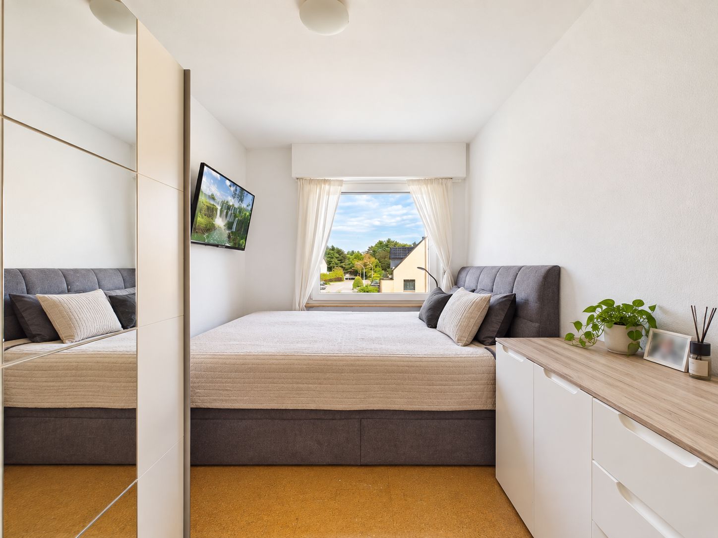Modernes Schlafzimmer mit großem Bett, Fenster, Spiegel und Kommode mit Pflanzen.
