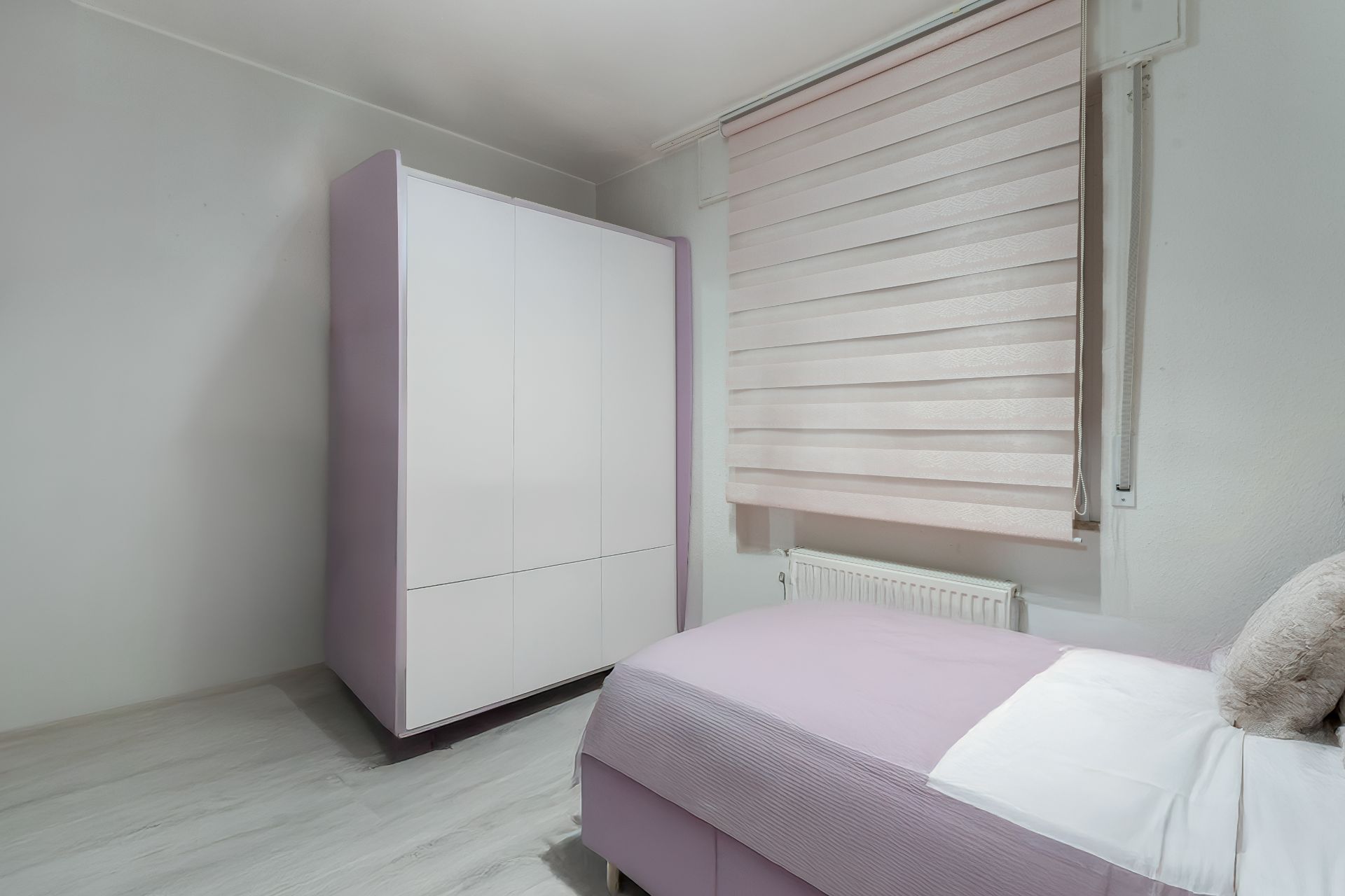 Helles Schlafzimmer mit lila Bett, weißem Kleiderschrank und Fenster mit Jalousie.