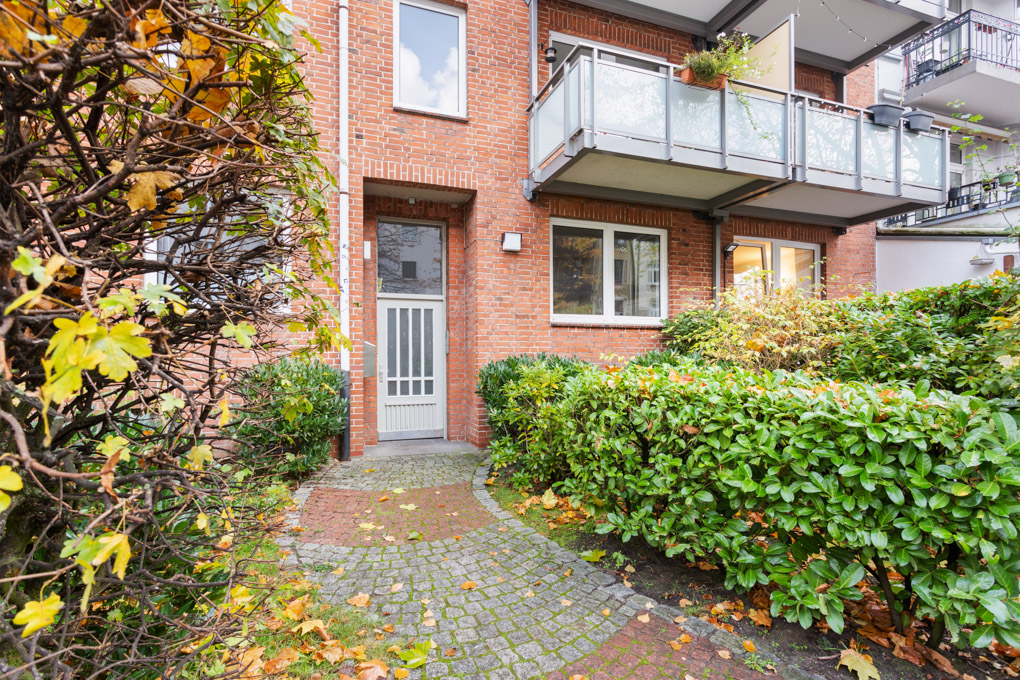 Eingang einer Wohnhauswohnung mit Ziegelmauer, umliegenden Büschen und Balkon.