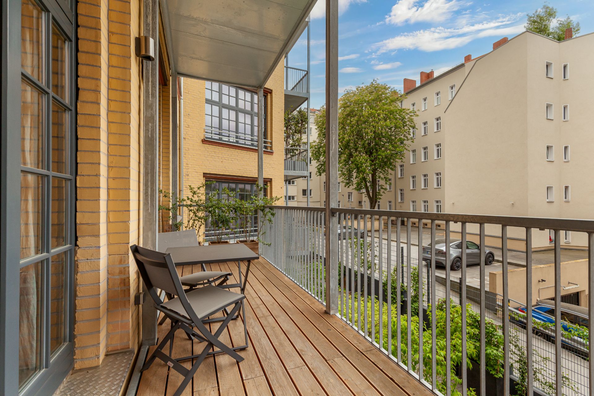Balkon mit Holzboden, Tisch und zwei Stühlen, Blick auf Wohnhäuser und Straße.