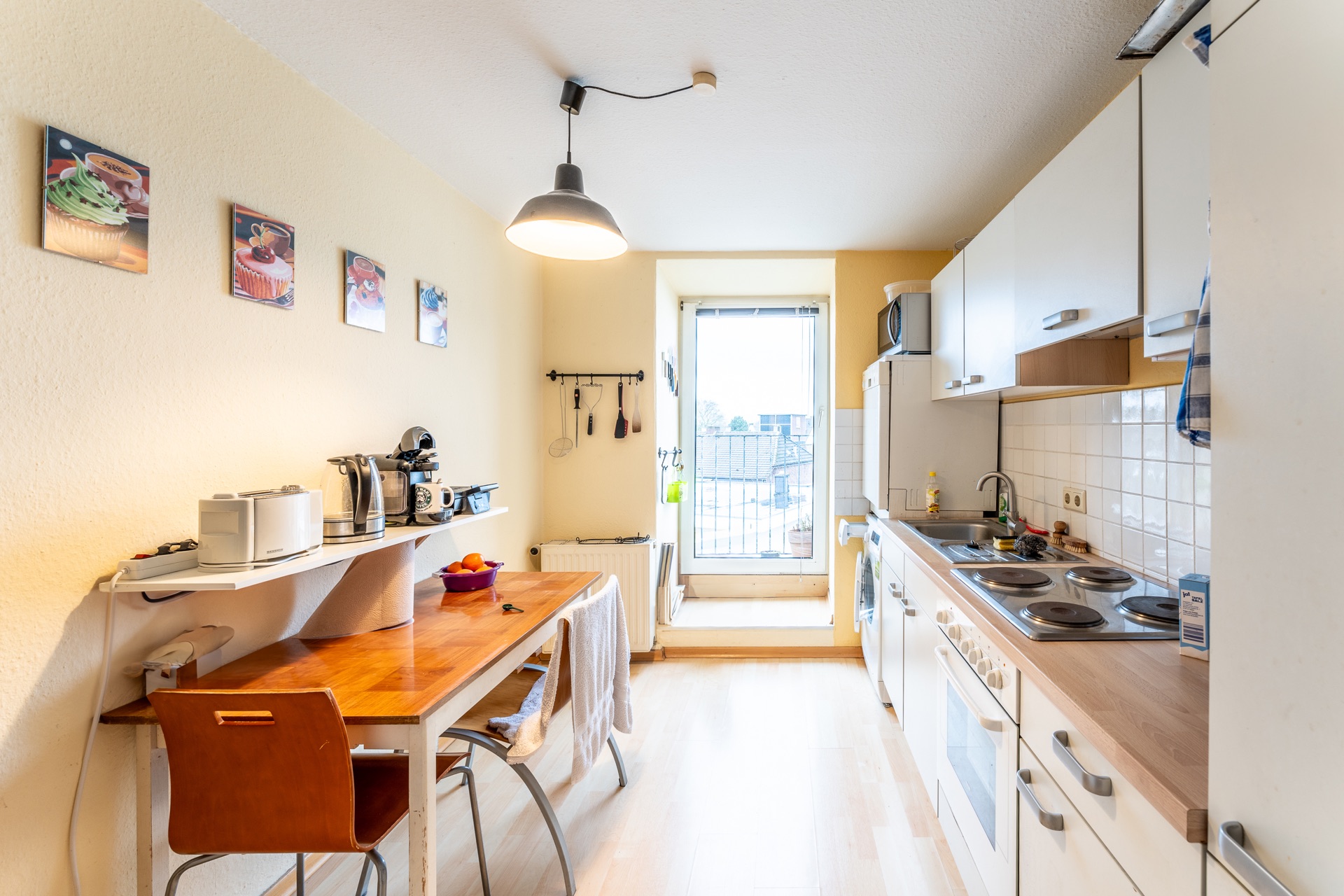 Helle Küche mit Holztisch, Stühlen, Einbauküche, großen Fenstern und Küchenutensilien an der Wand.