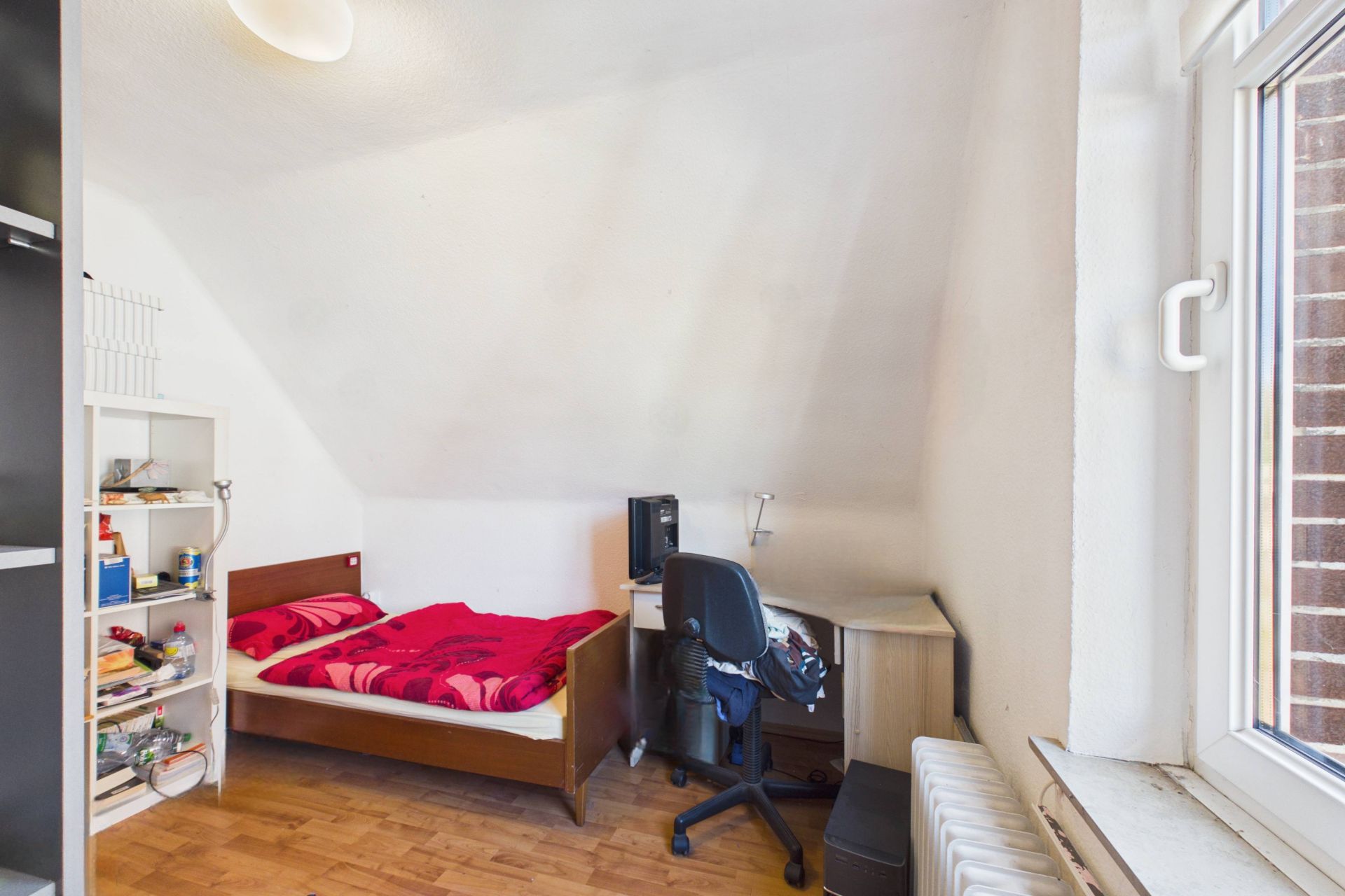Kleines Zimmer mit Einzelbett, Schreibtisch, Stuhl und Regal, Holzboden, Fenster rechts.