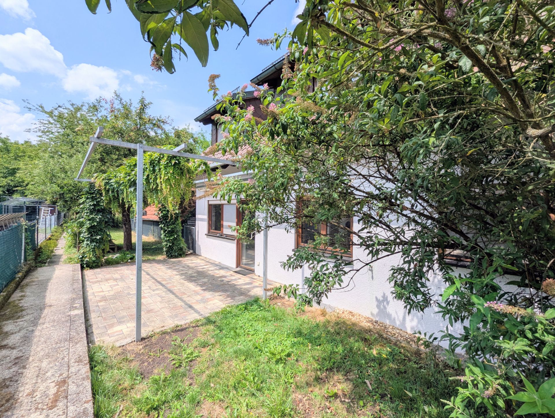 Garten mit Terrasse und Weg neben einem Wohnhaus, Bäume und Büsche im Vordergrund.