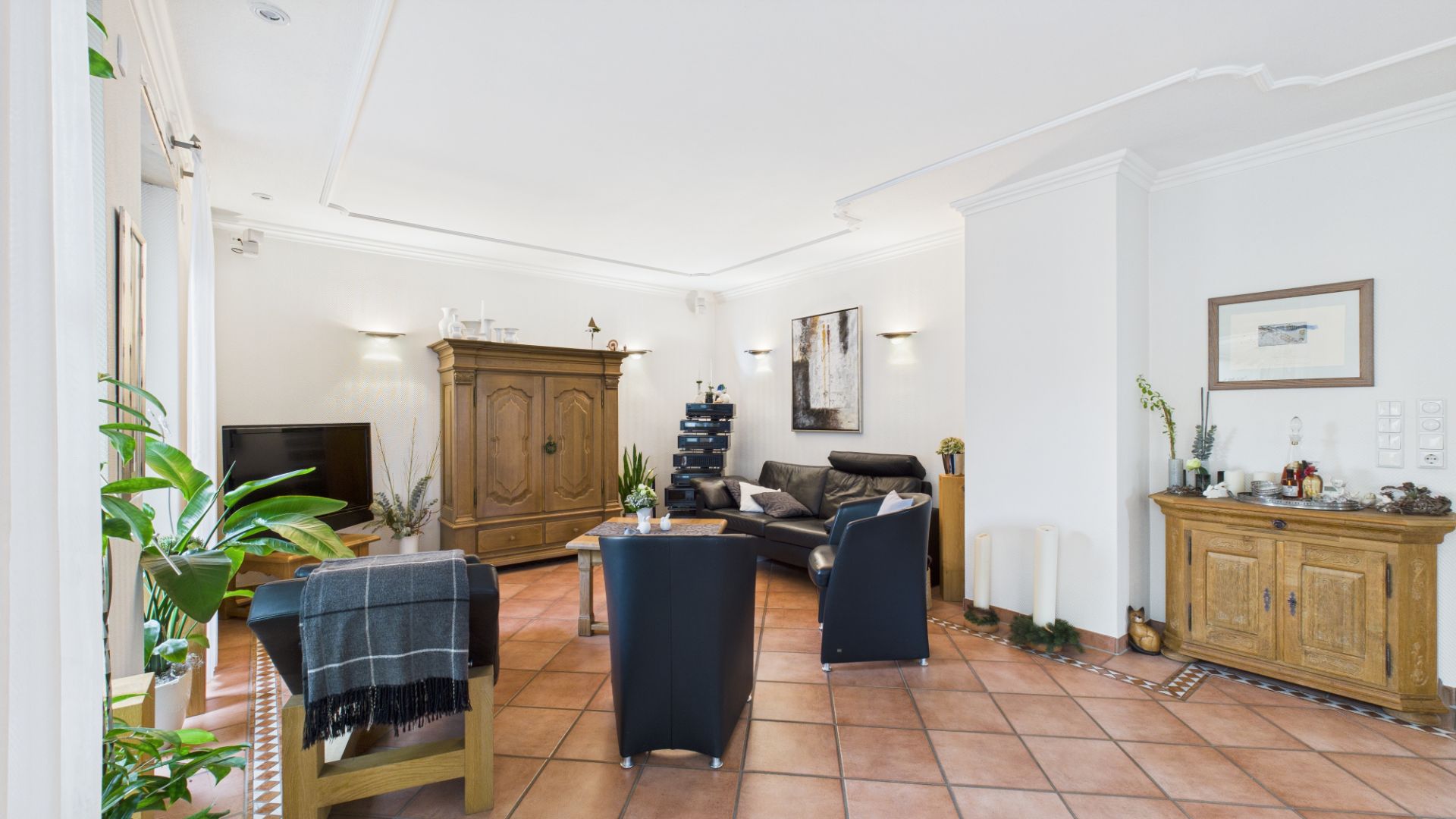 Helles Wohnzimmer mit Sofa, Sesseln, Holzmöbeln, Pflanzen und Fliesenboden.