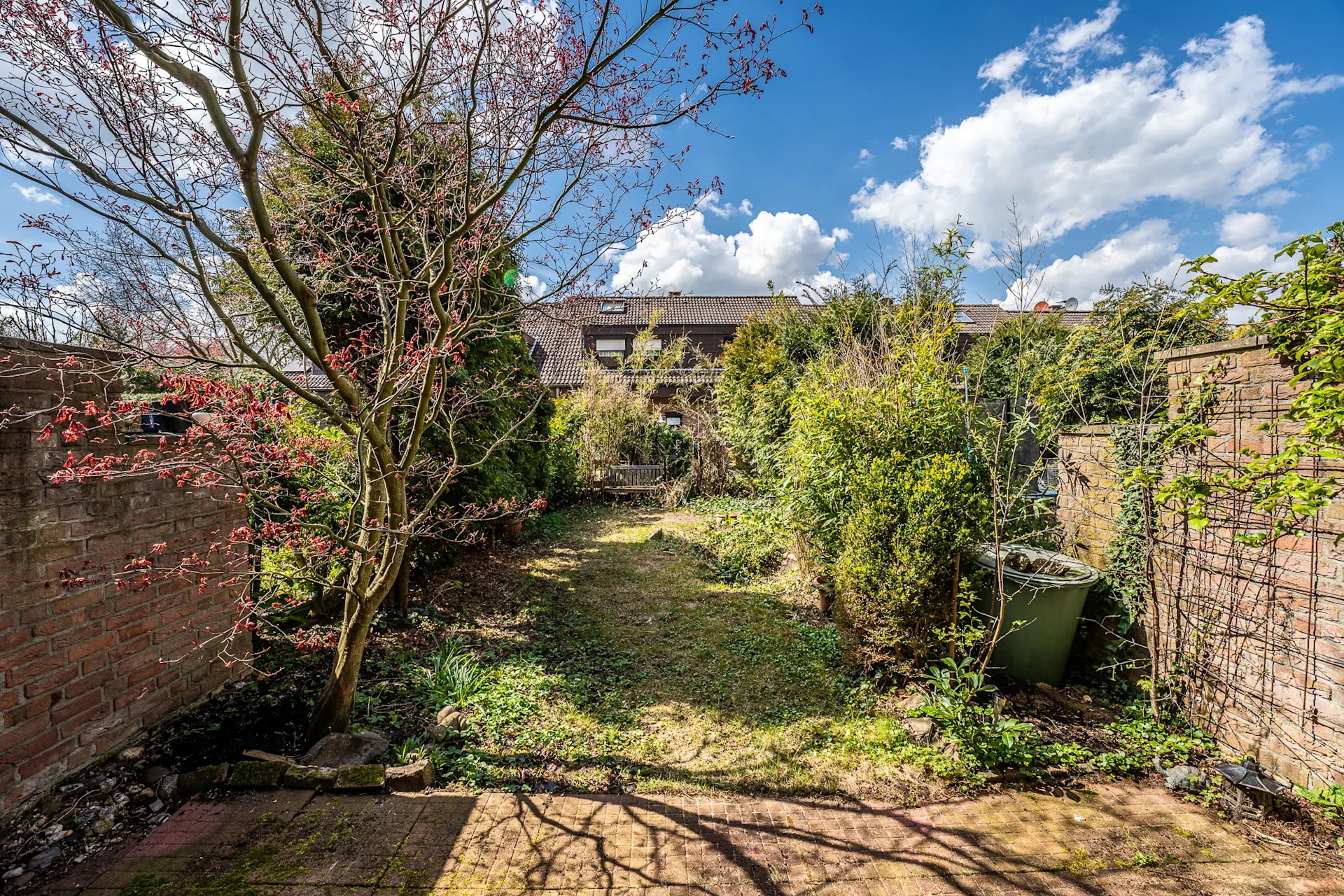 Kleiner Garten mit Baum, Sträuchern, Rasen und einer Mülltonne neben einer Backsteinmauer.
