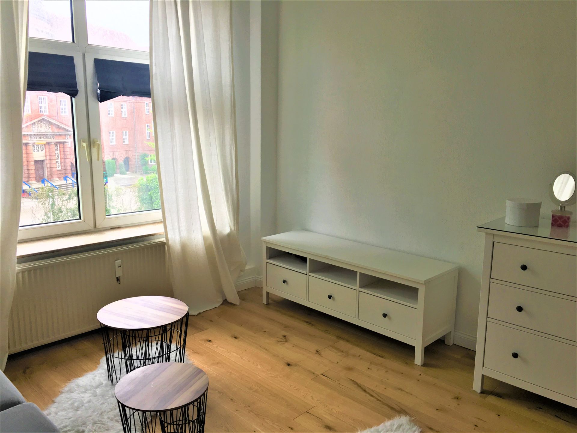 Zimmer mit großem Fenster, weißer Kommode, niedrigen Tischen und Vorhängen auf Holzfußboden.