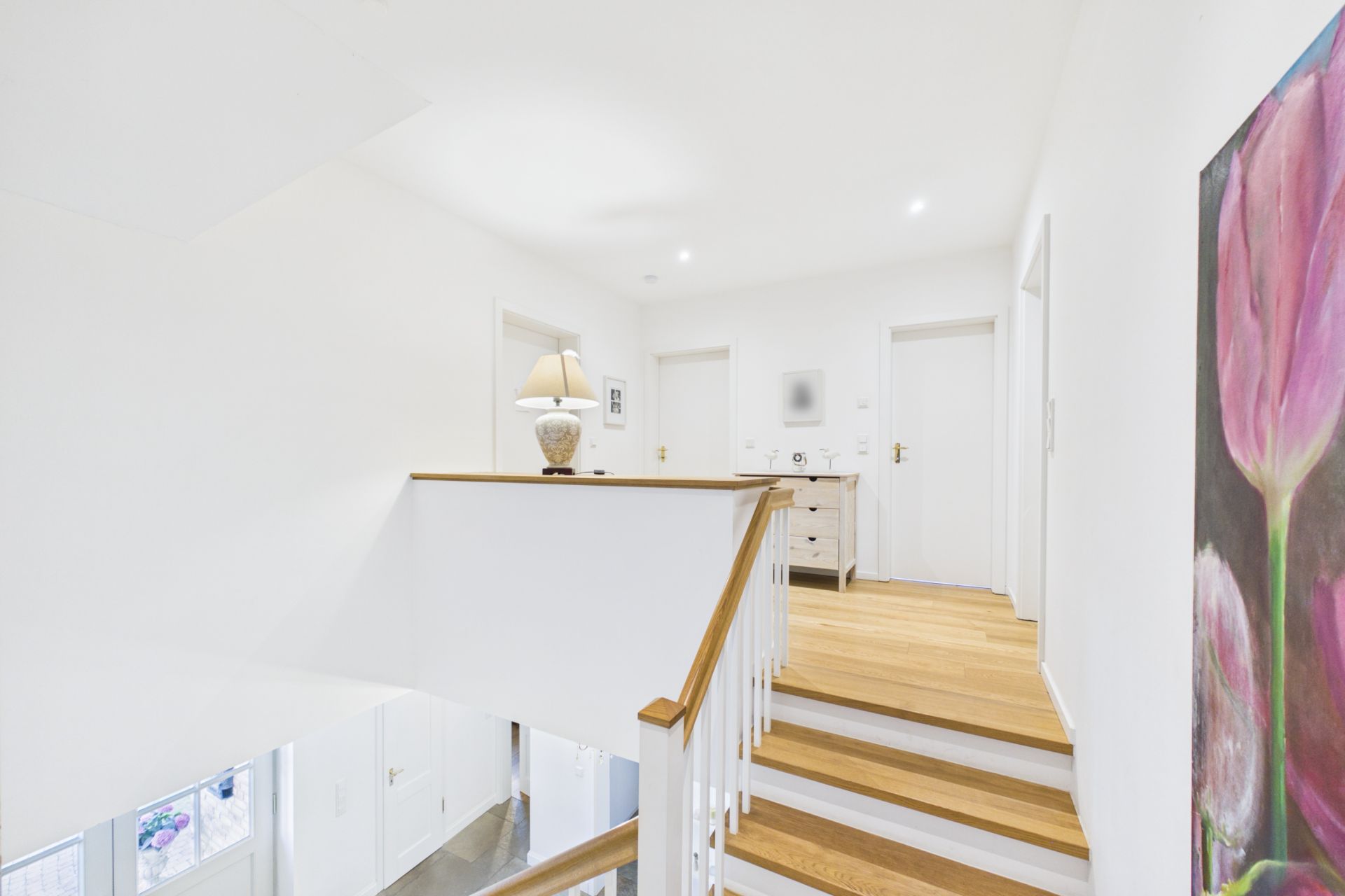 Heller Flur mit Holztreppe, Kommode, Lampe und großem Blumenbild an der Wand.
