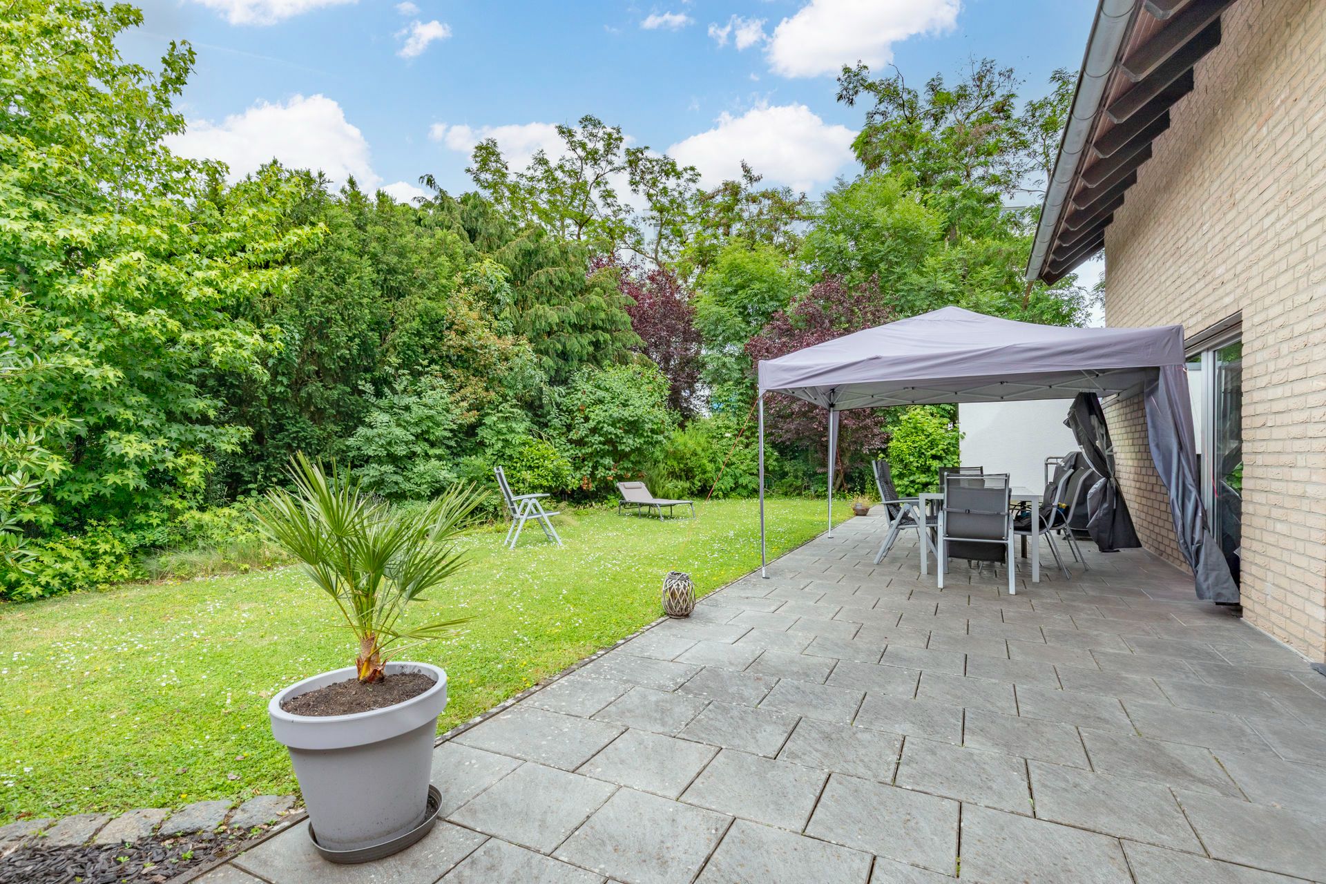 Gepflasterte Terrasse mit Pavillon, Gartenmöbeln und Topfpflanze, angrenzend an einen grünen Garten.