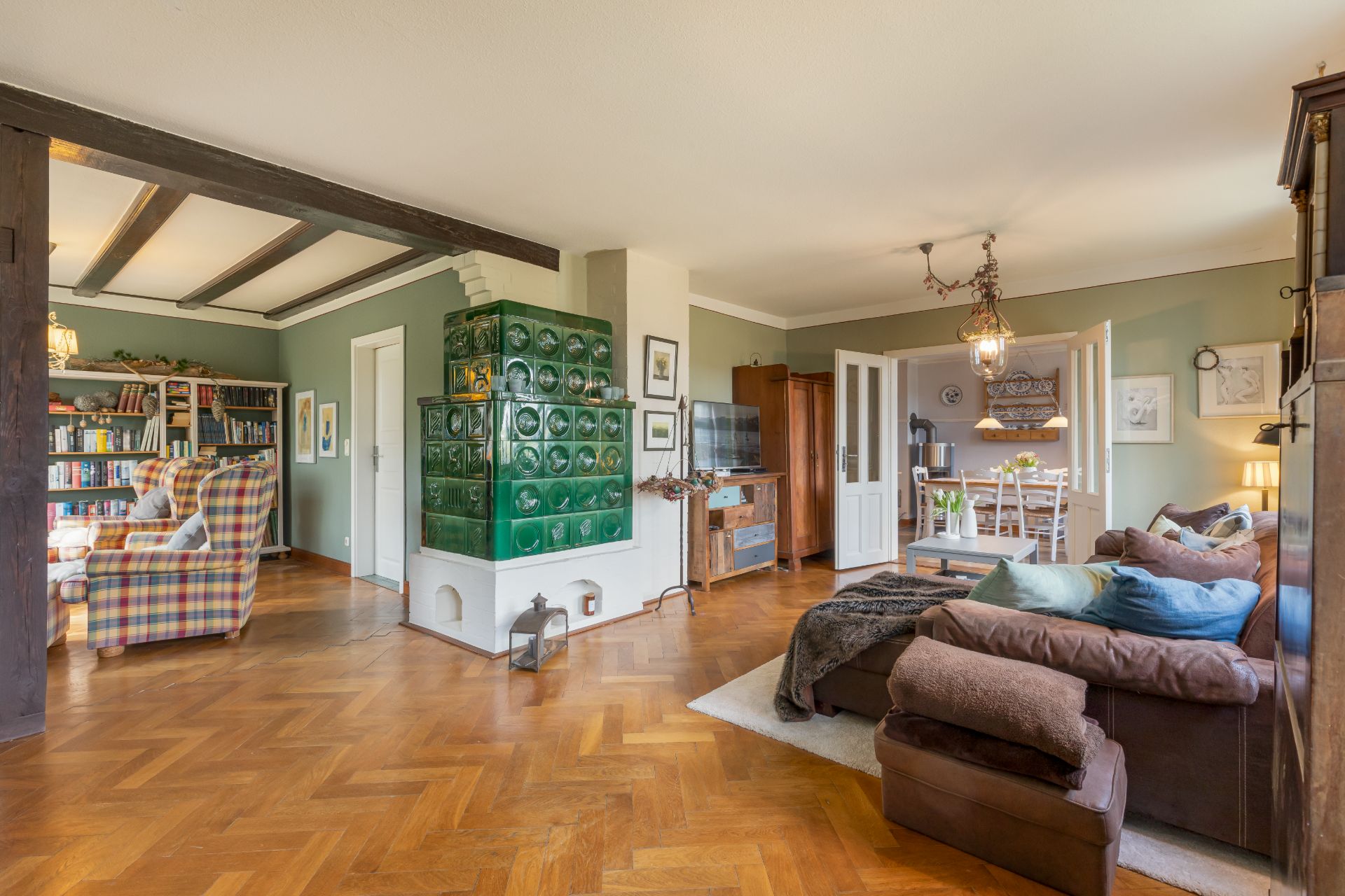 Wohnzimmer mit grünem Kachelofen, Holzboden, Sofa und angrenzendem Essbereich.