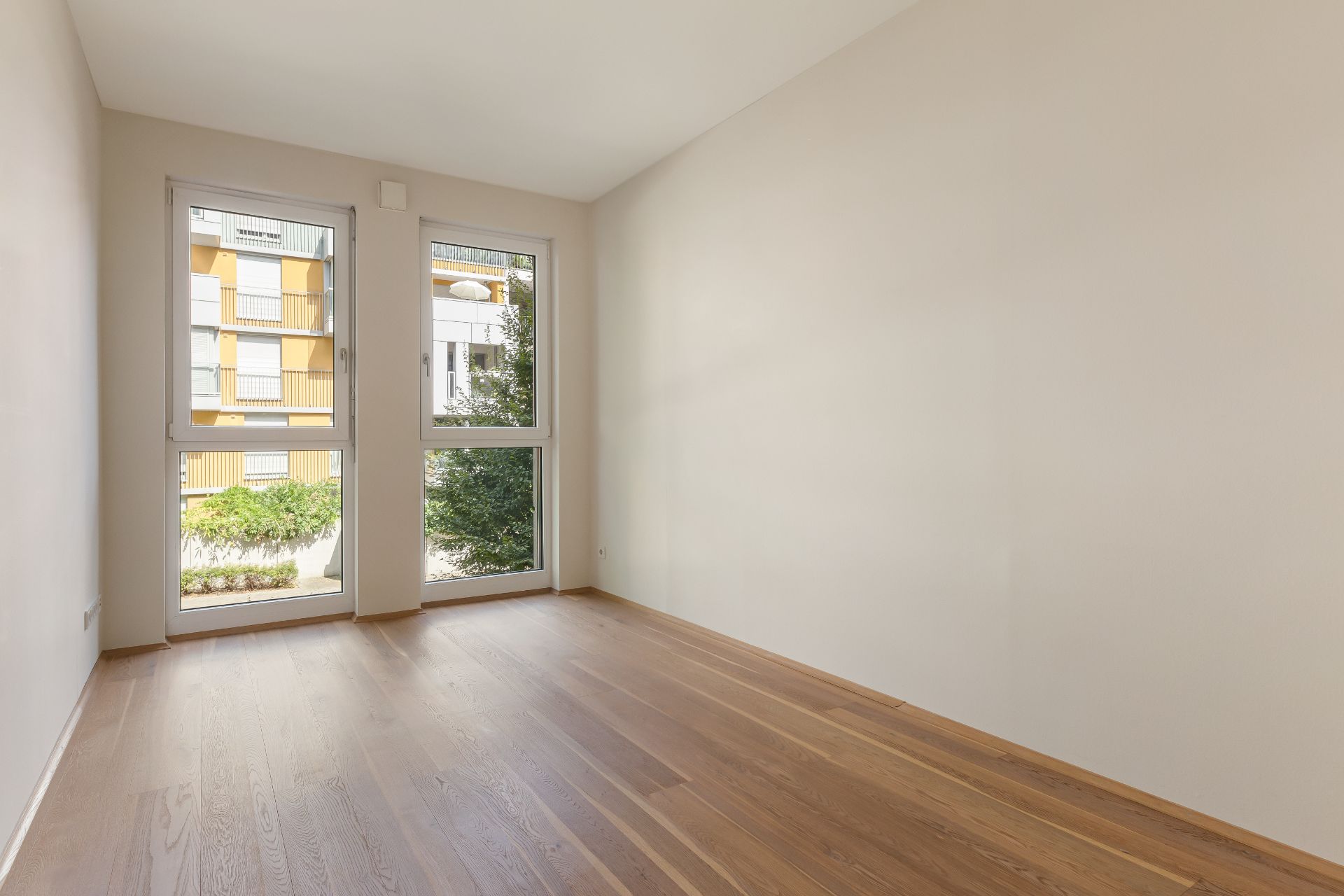 Leerer, heller Raum mit Holzboden und großen Fenstern, Blick auf Außenbereich mit Bäumen.