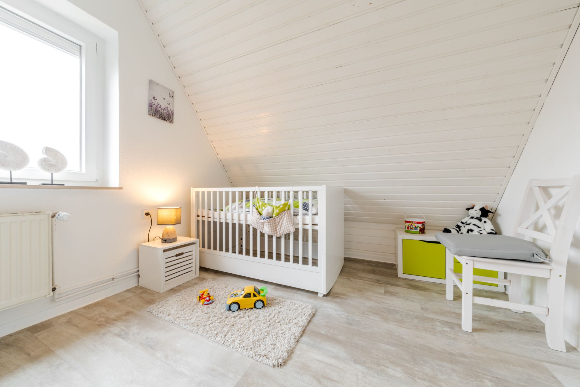 Helles Kinderzimmer mit Gitterbett, Teppich, Spielzeugauto und Stuhl unter einer Schräge.