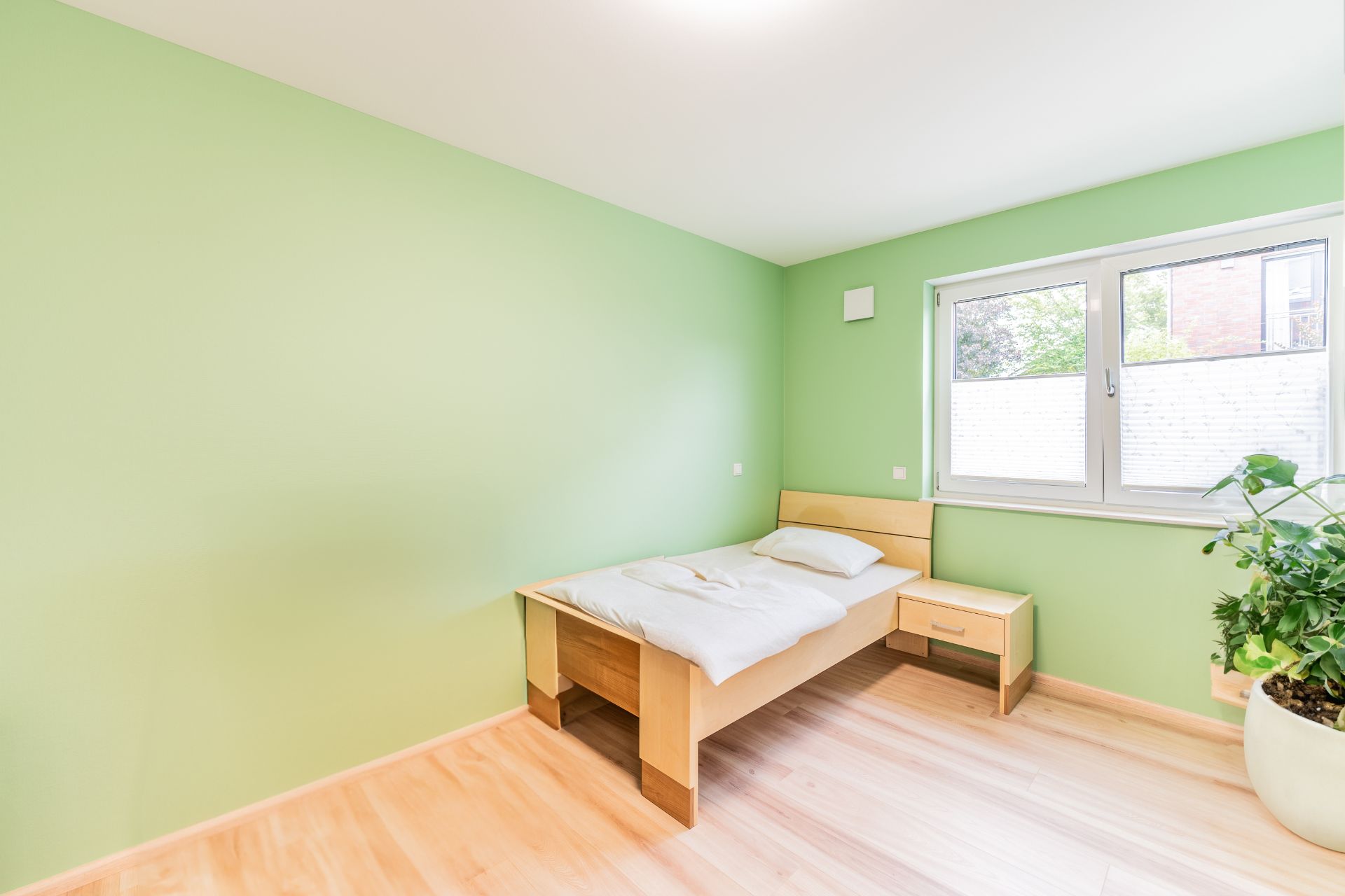 Helles Schlafzimmer mit grün gestrichenen Wänden, Einzelbett, Nachttisch und großem Fenster.
