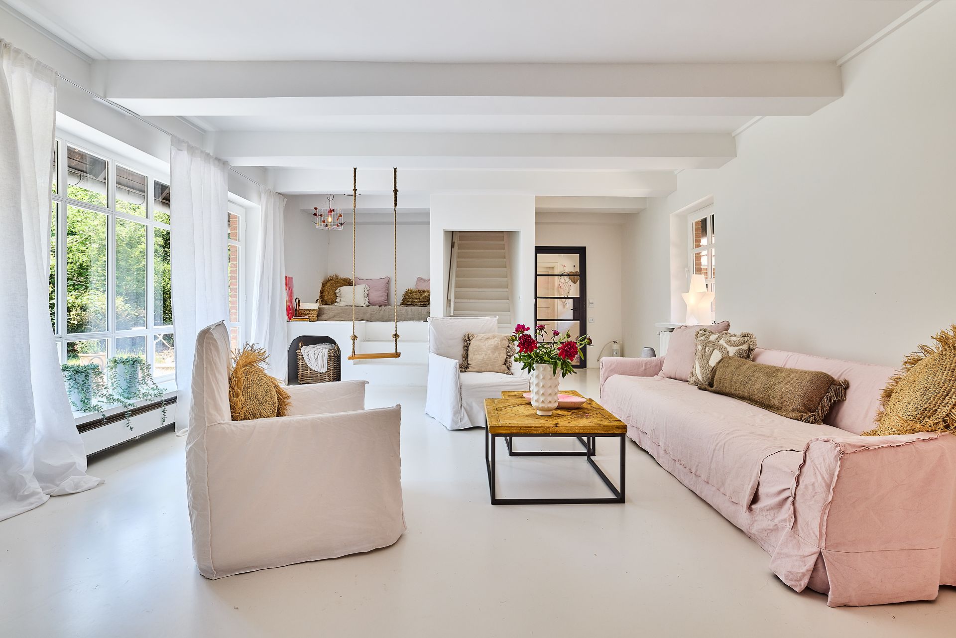 Helles Wohnzimmer mit großen Fenstern, rosa Sofa, Sessel, Holztisch und Schaukel.