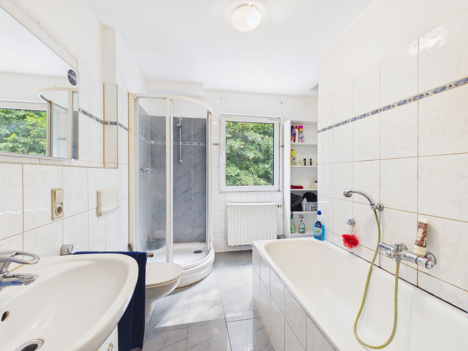 Helles Badezimmer mit Dusche, Badewanne, Waschbecken und Fenster mit Blick ins Grüne.