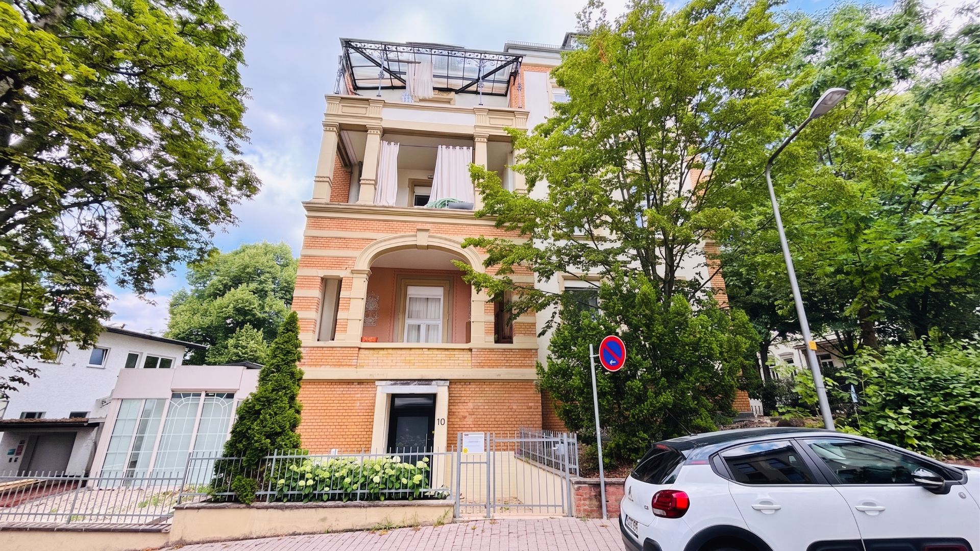Mehrstöckiges Wohngebäude mit Balkon, Bäumen davor und geparktem Auto auf der Straße.