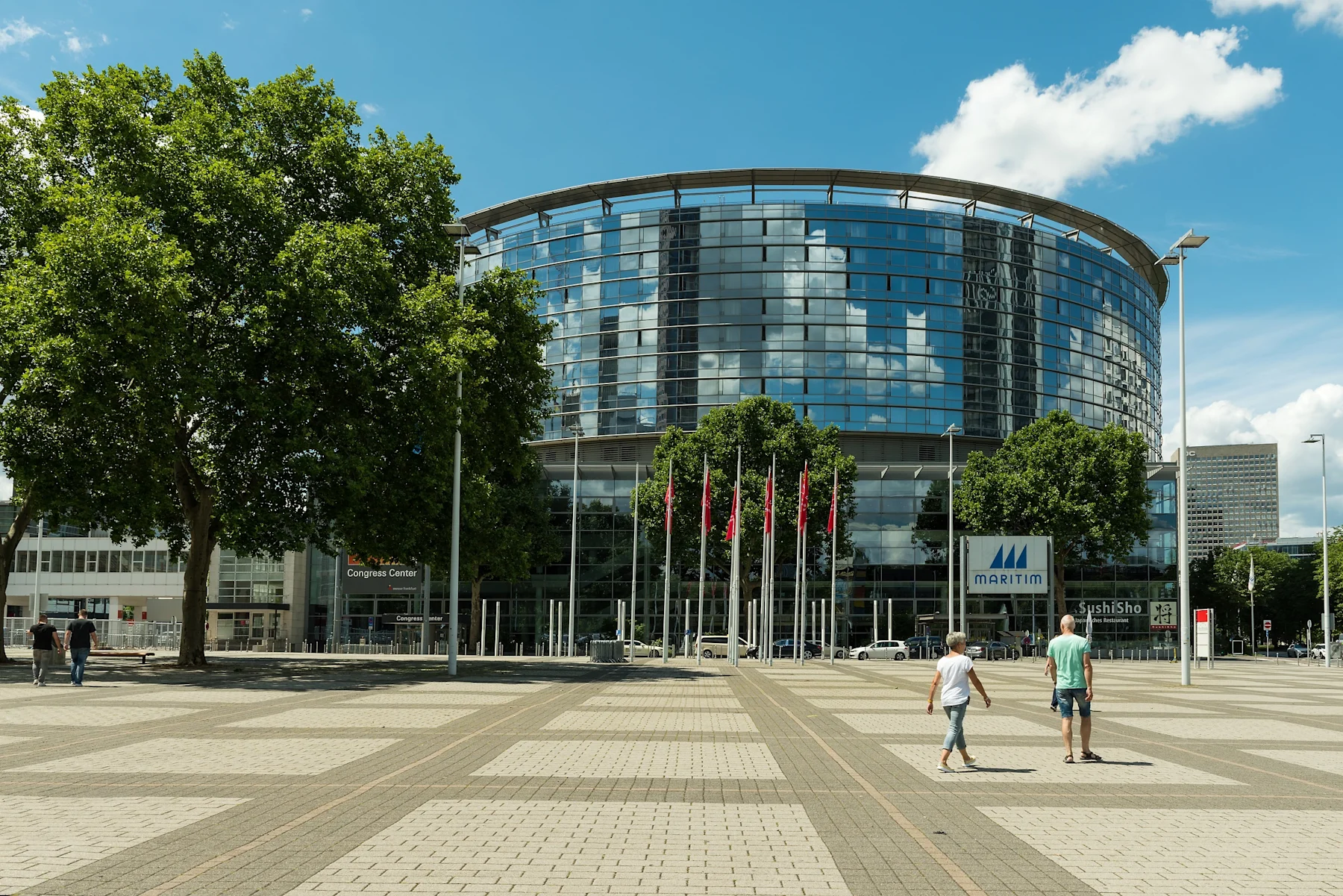 Glasgebäude mit Bäumen und mehreren roten Fahnen im Vordergrund auf einem Platz.