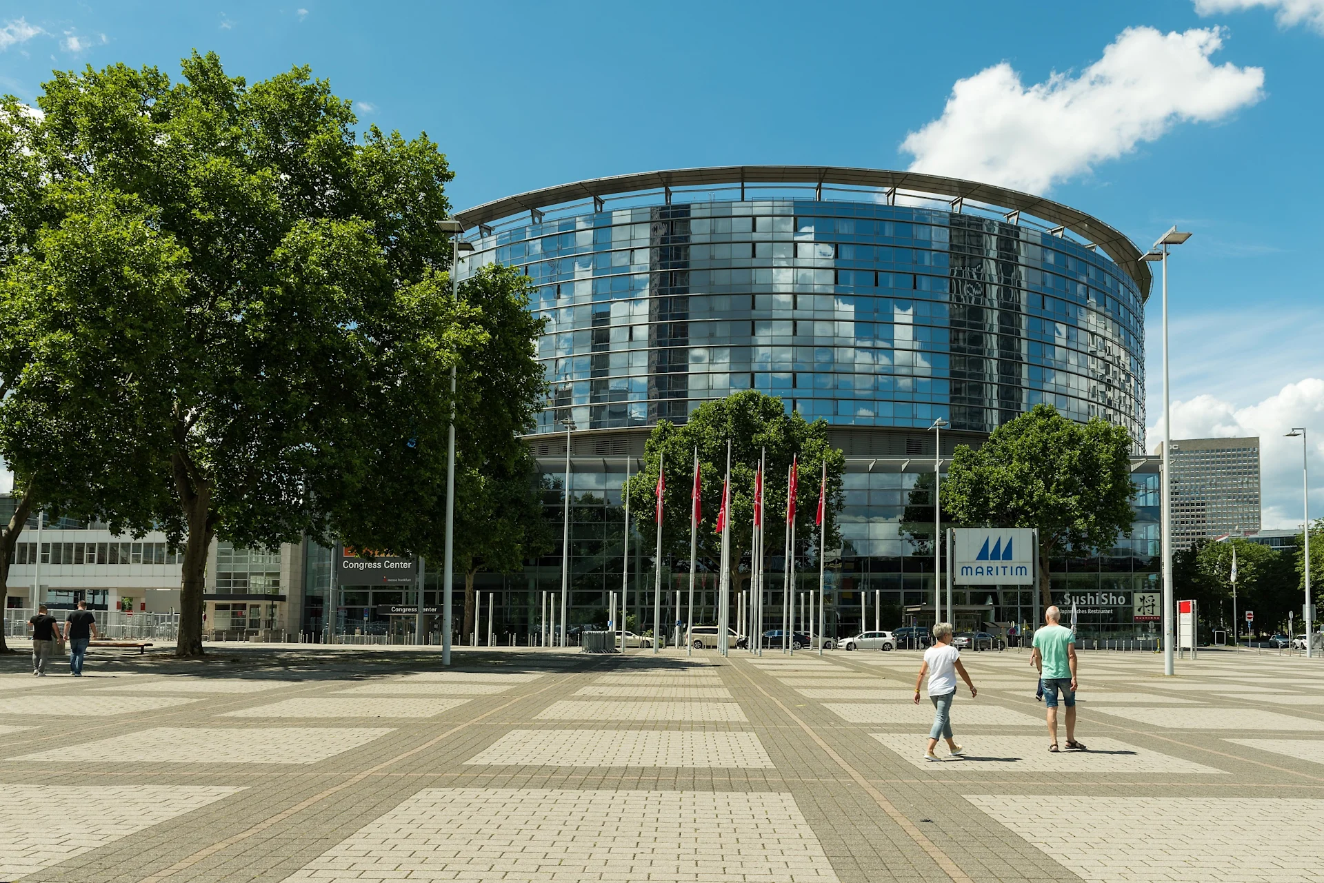 Glasgebäude mit Bäumen und mehreren roten Fahnen im Vordergrund auf einem Platz.