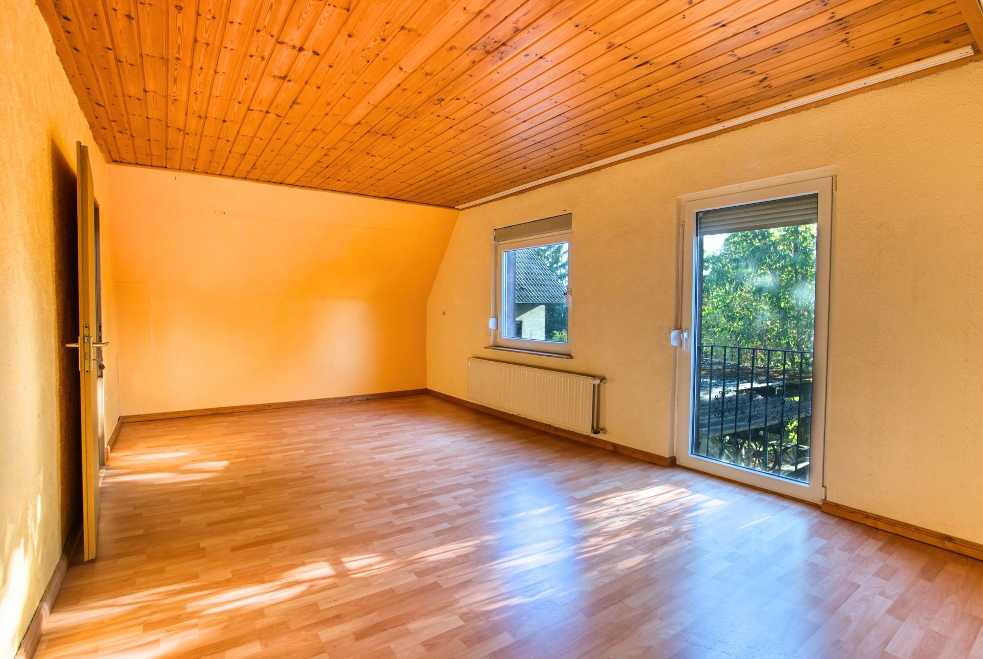 Helles, leeres Zimmer mit Holzdecke, Laminatboden, Fenster und Tür zum Balkon.