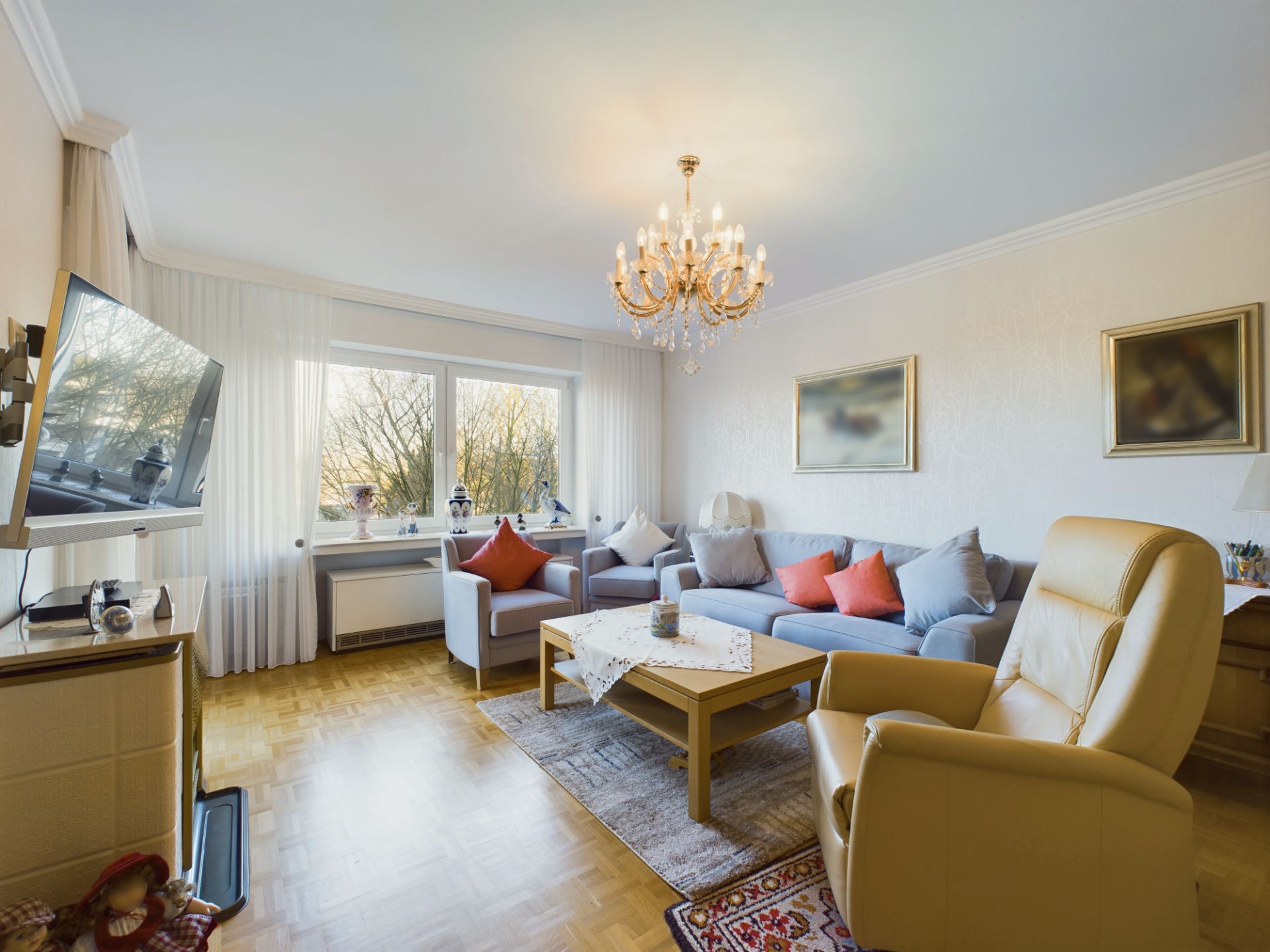 Helles Wohnzimmer mit Sofa, Sessel, Couchtisch, großem Fenster und Kronleuchter.