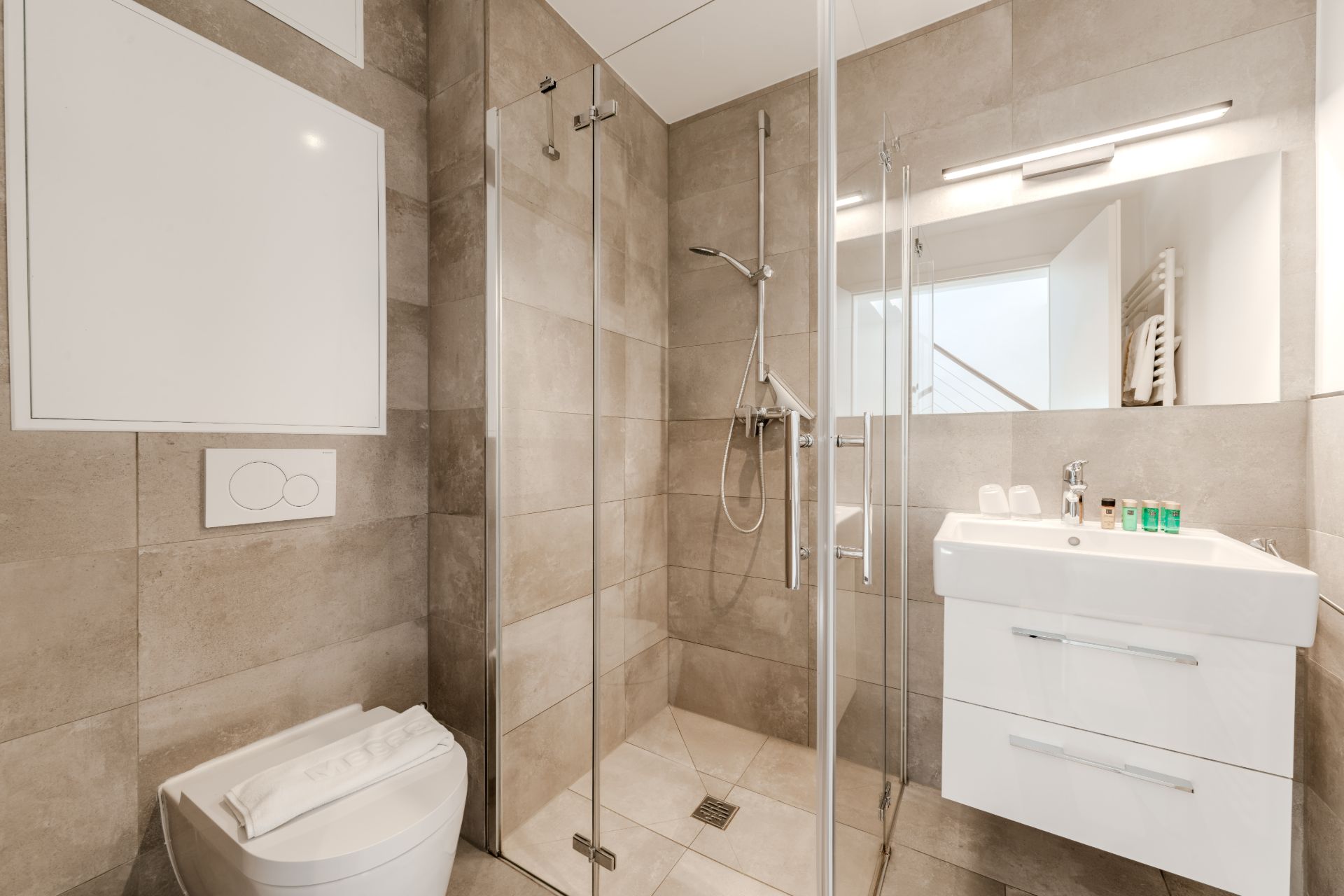 Modernes Bad mit großer Dusche, Wandtoilette, Waschtisch mit Spiegel und hellen Fliesen.